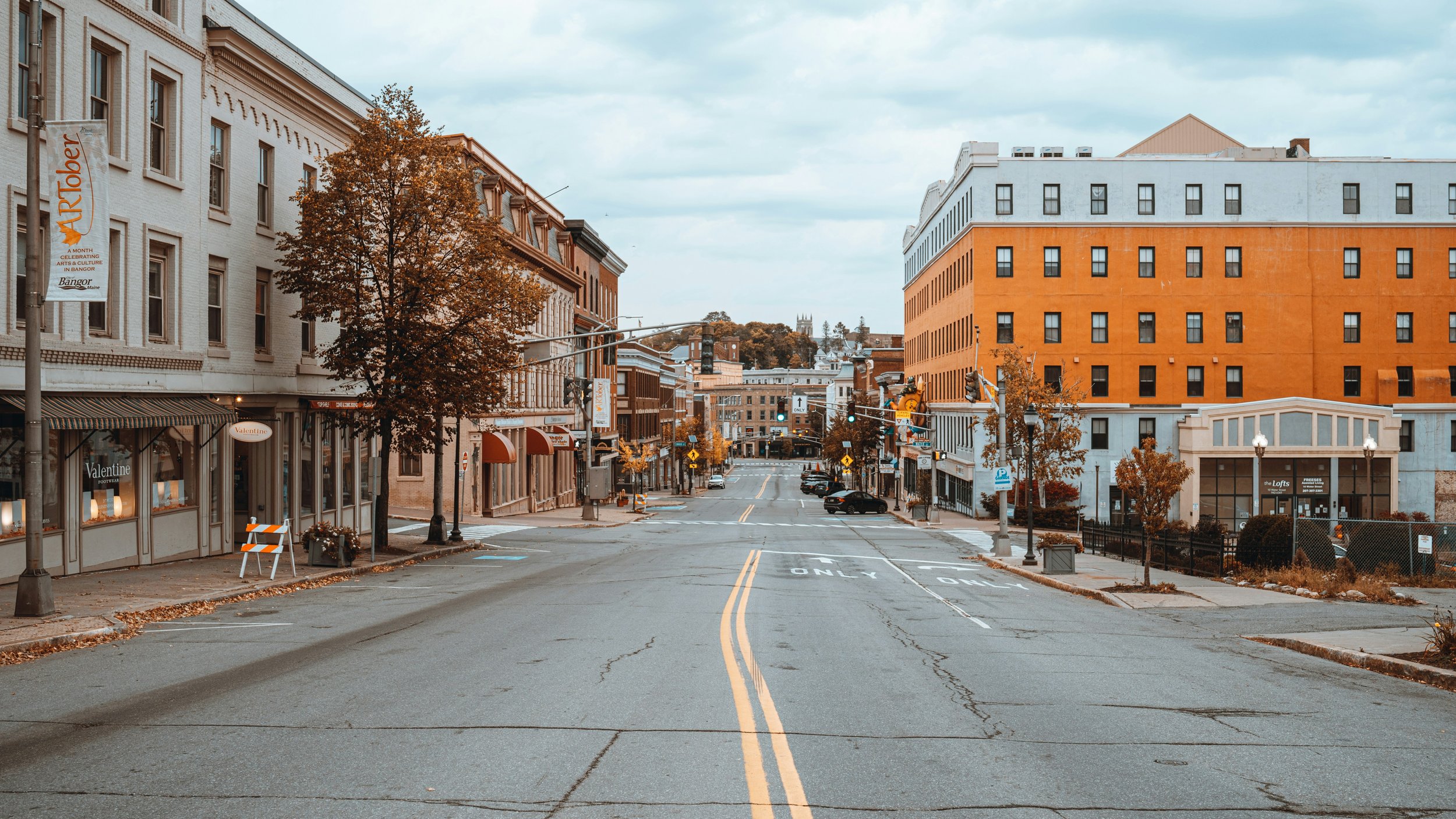 Empty city street with buildings on both sides, some trees with autumn foliage, and traffic lights at the intersection.