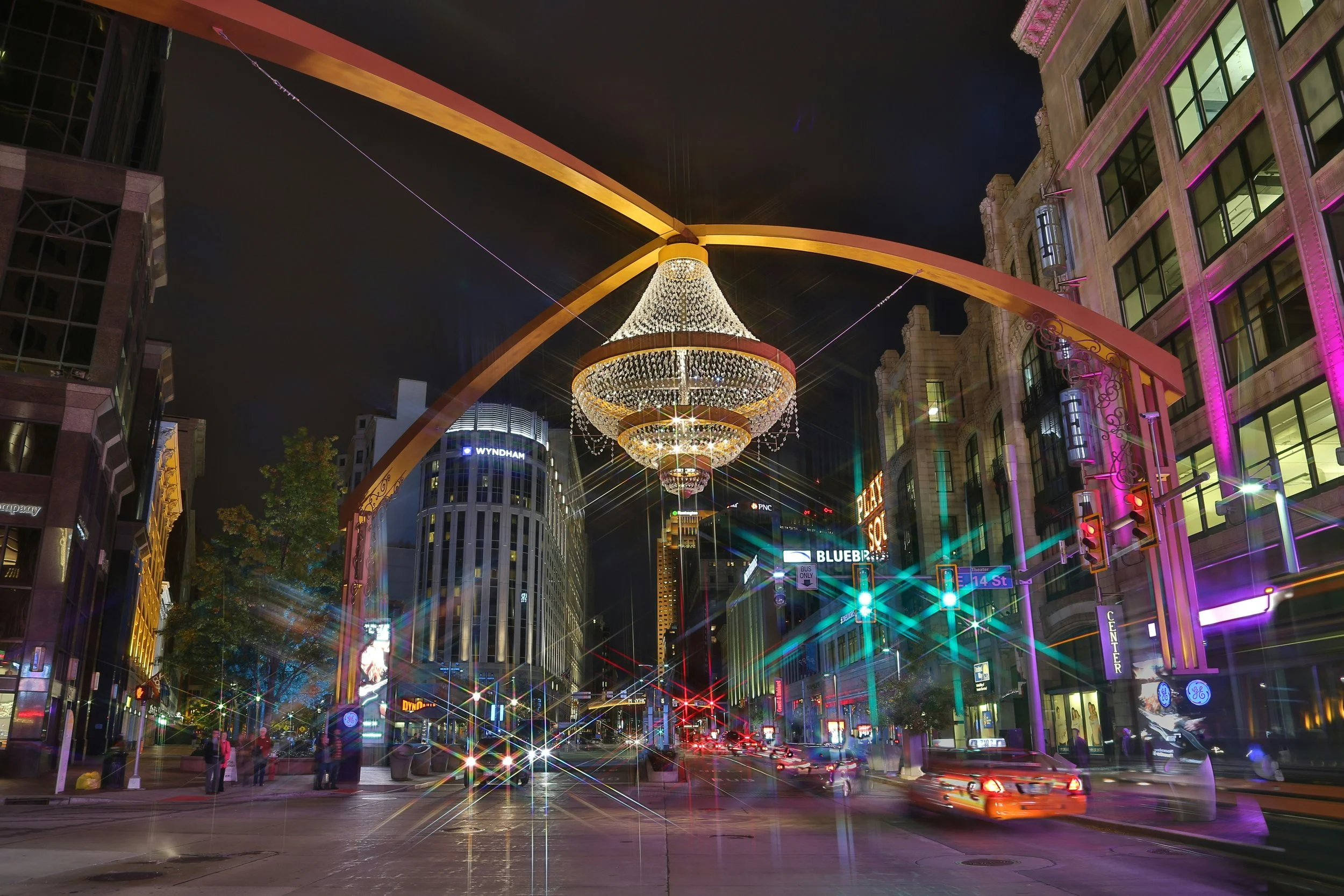 Nighttime city street scene with illuminated buildings, colorful lights, and a large chandelier overhead strung across the street, with blurred moving cars and pedestrians.