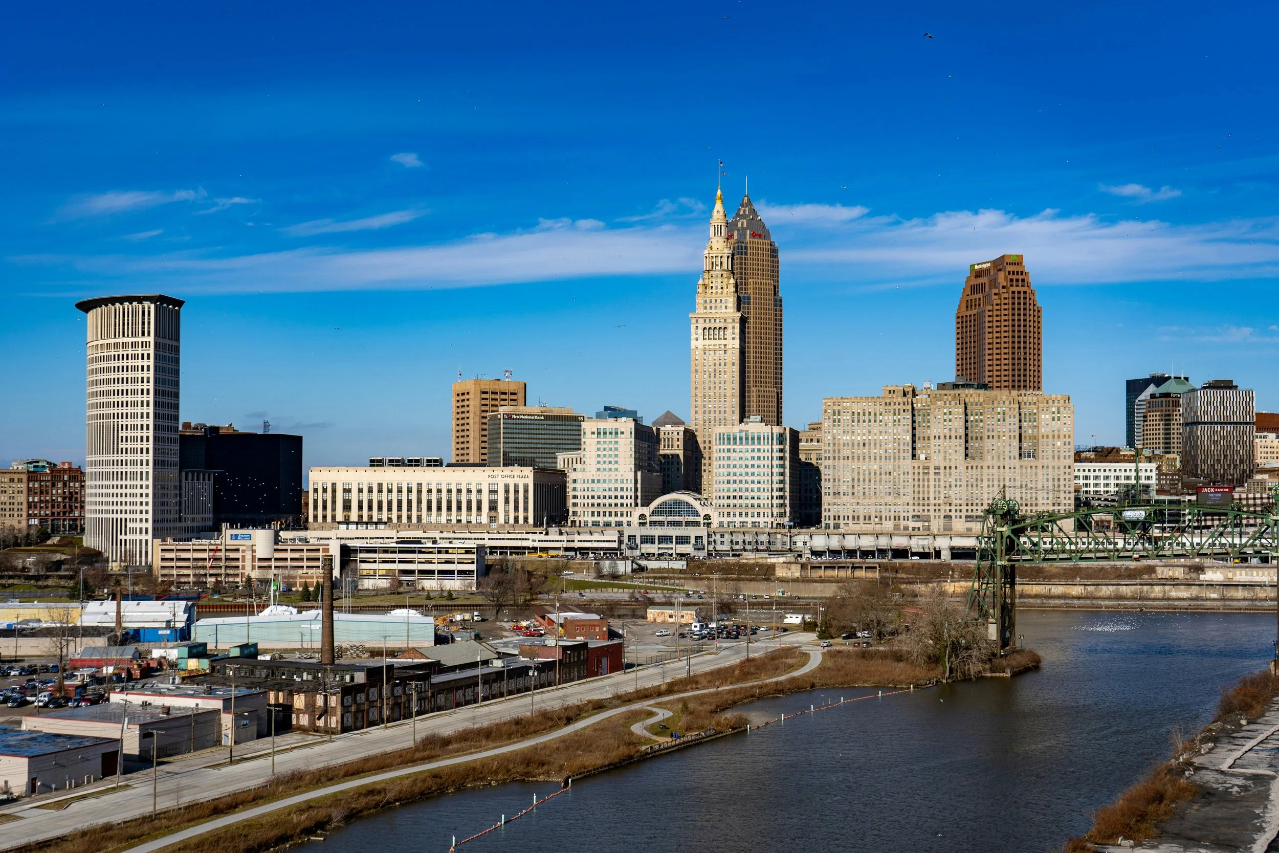 Skyline of Cleveland, Ohio with tall buildings including the Key Tower and Cleveland Trust Building, seen across a river with a park and paths in the foreground.
