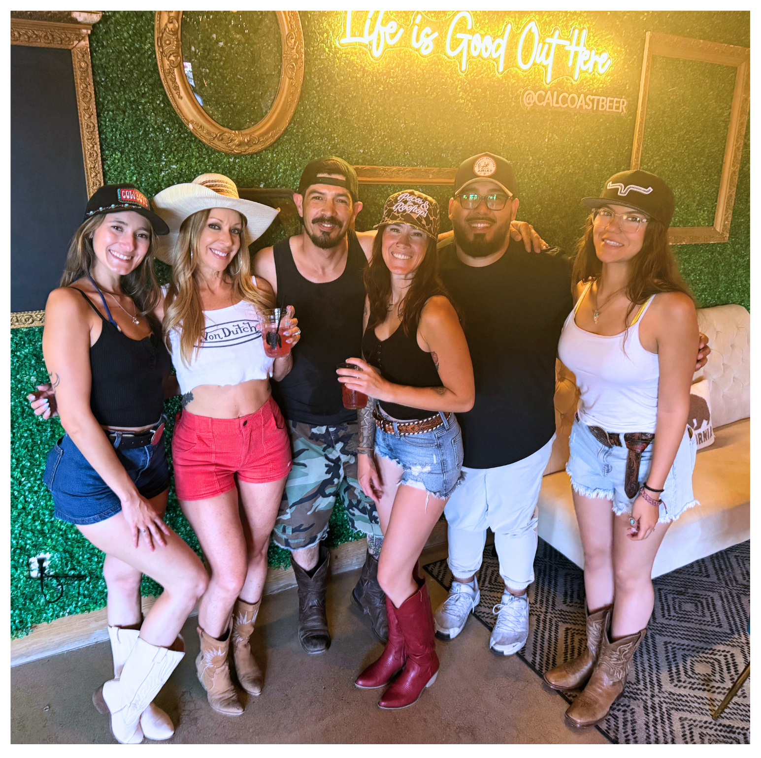 Group of six friends posing indoors in front of a green foliage wall with a neon sign that reads "Life is Good Out Here" and an Instagram handle '@CALCOASTBEER'. They are dressed casually in summer outfits and cowboy boots, holding drinks, with some smiling and wearing hats or caps.