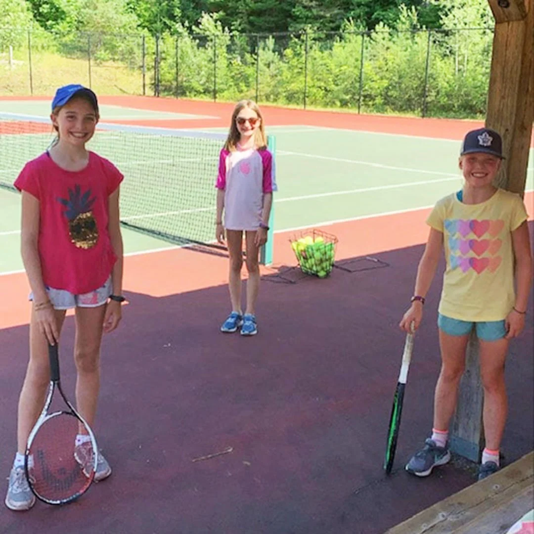 Three young girls on a tennis court with tennis rackets, smiling at the camera. There's a basket of tennis balls nearby, and the court is outdoors with green trees in the background.