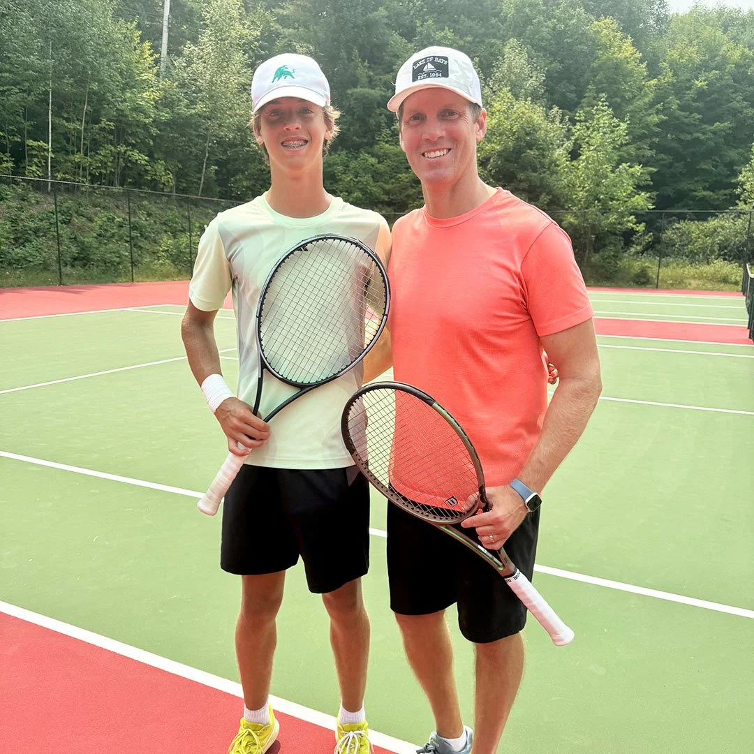 A boy and a man standing on a tennis court holding tennis rackets and smiling at the camera, dressed in athletic clothing and hats.