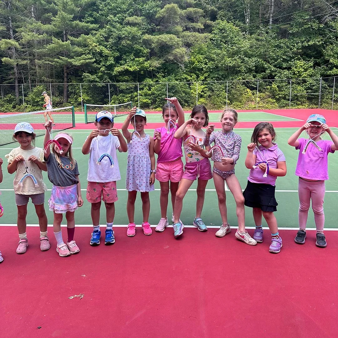 Group of young children standing in a row on a tennis court, holding rainbow-shaped crafts, with trees in the background.