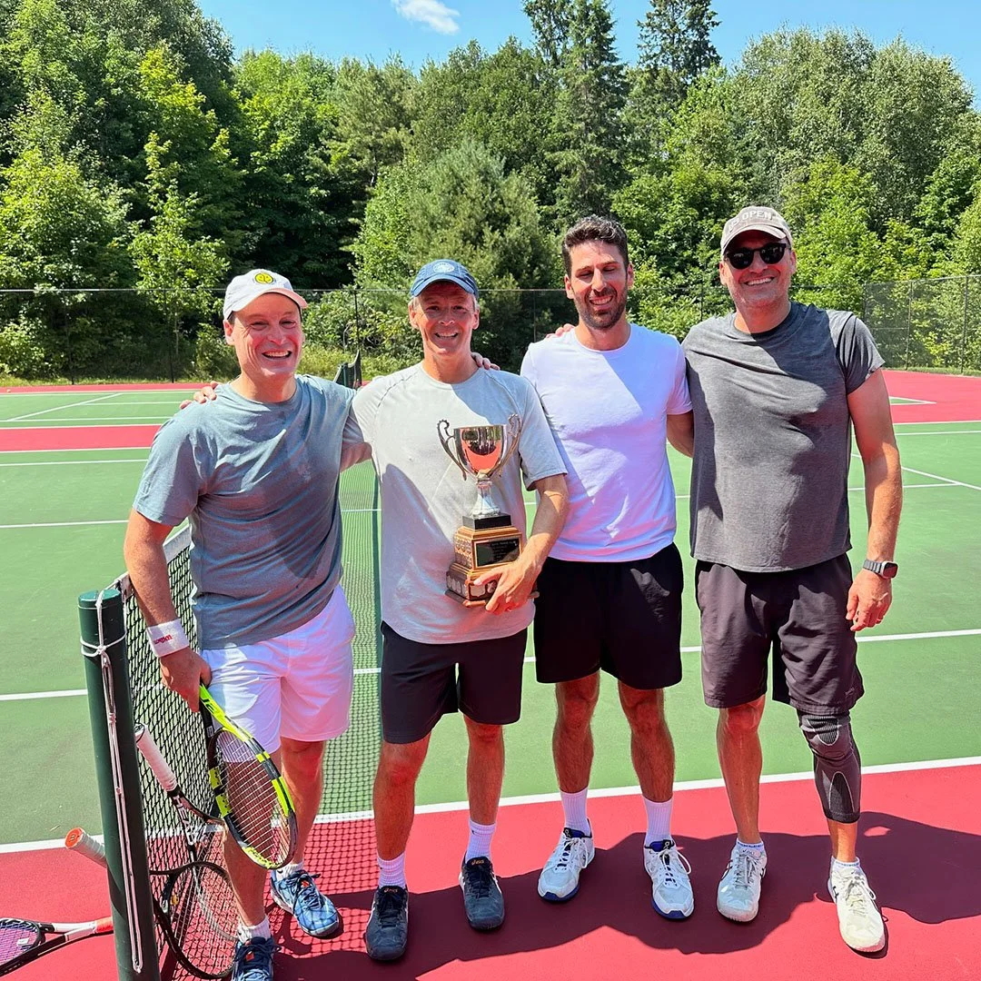 Four men standing on a tennis court, smiling and holding a trophy, with trees in the background.