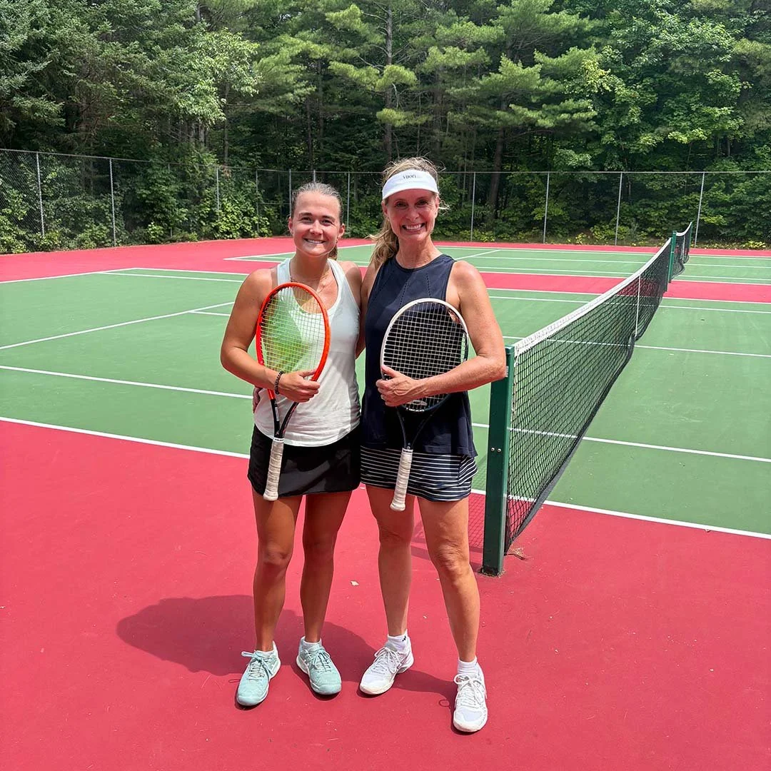 Two women standing on a tennis court, smiling and holding tennis rackets, with a net between them, surrounded by trees.