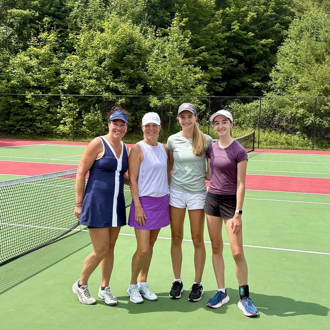 Four women standing together on a tennis court, dressed in sportswear and hats, with green trees in the background.