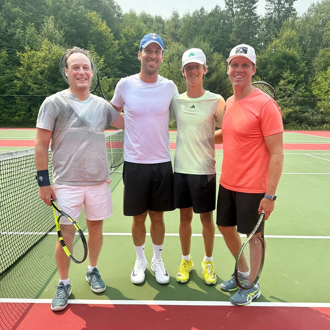 Four people standing on a tennis court, holding tennis rackets, smiling. They are dressed in athletic clothes, with trees and a chain-link fence in the background.