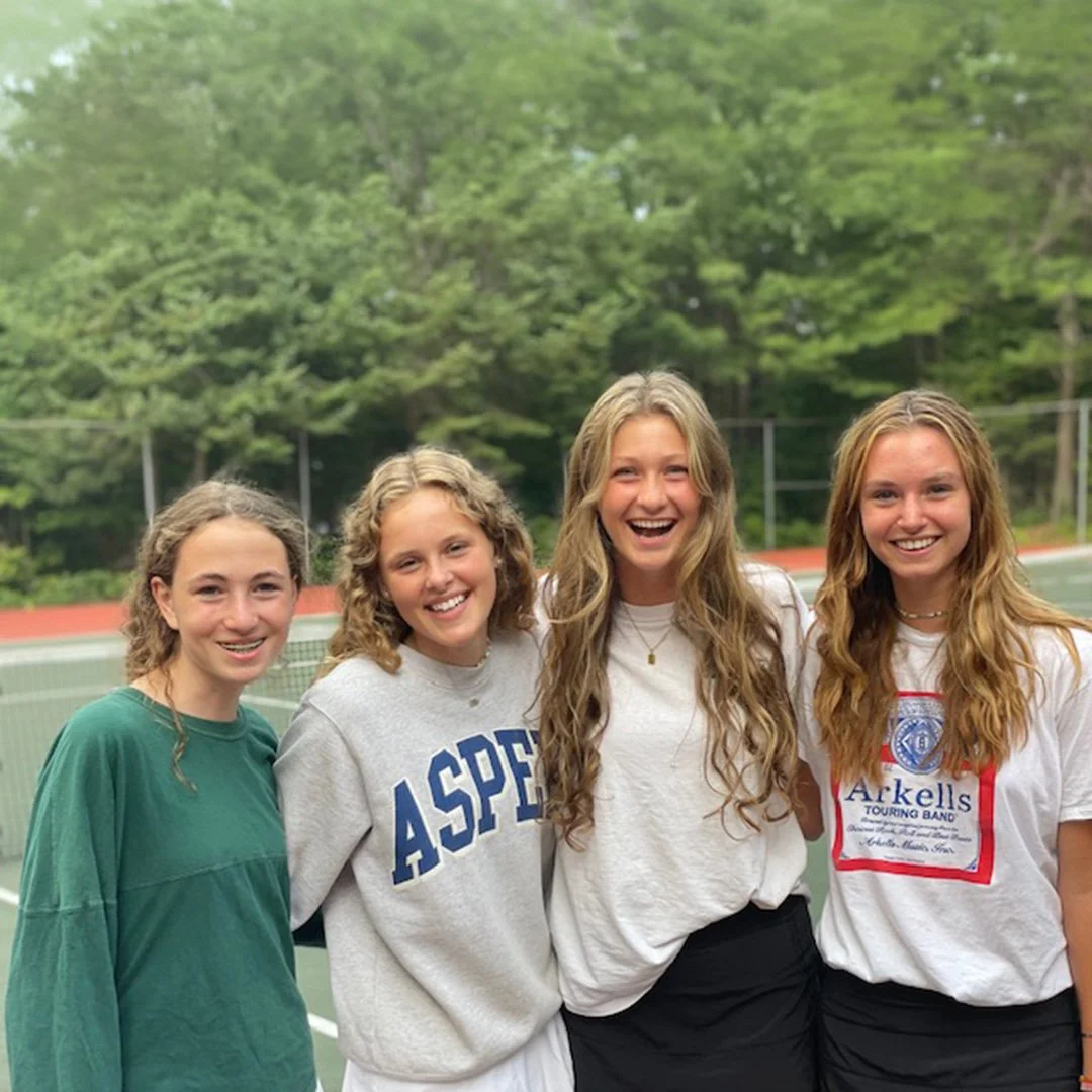 Four young women standing together on a tennis court, smiling, with trees in the background.