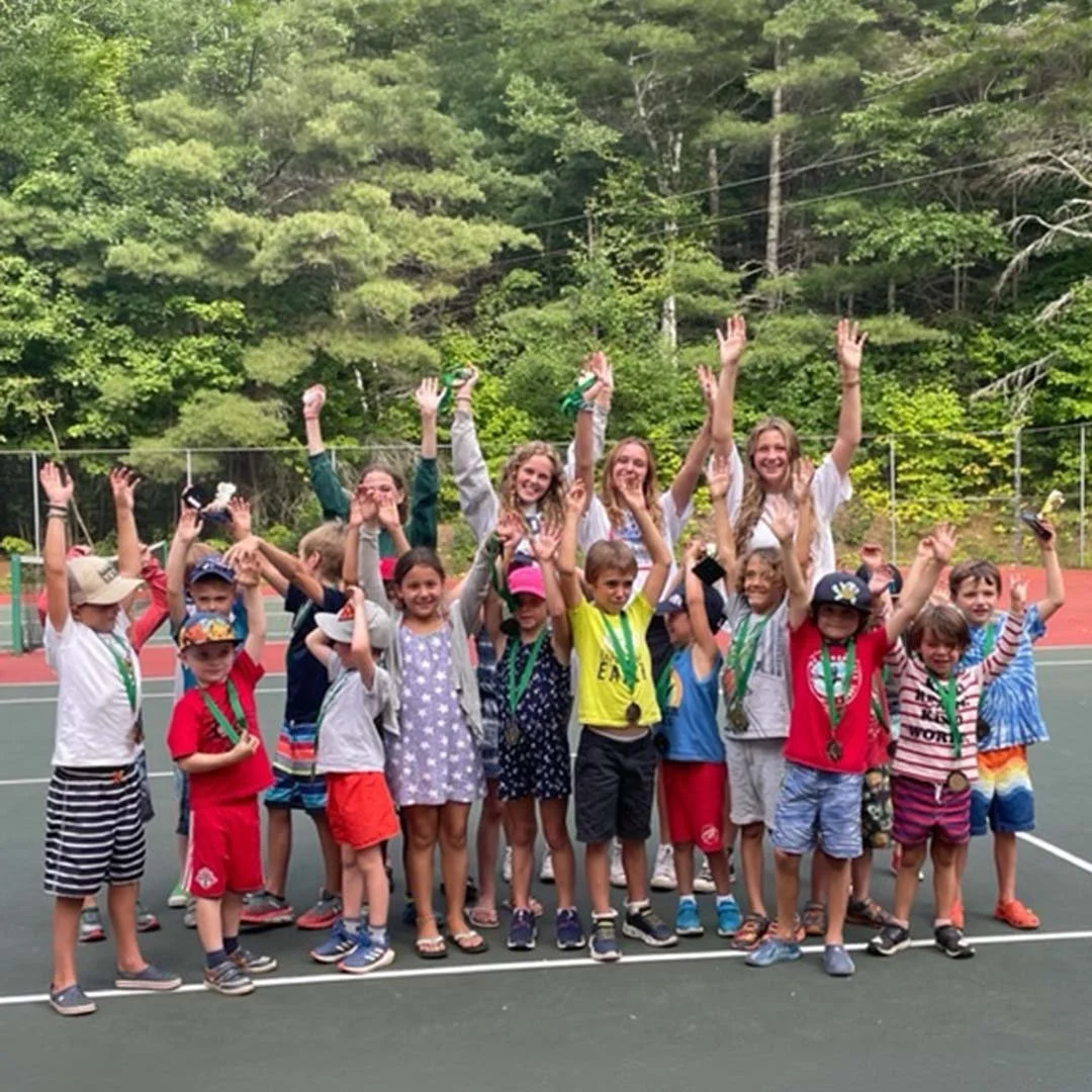 Group of children and young women celebrating on a tennis court, some wearing medals, with a forested background.