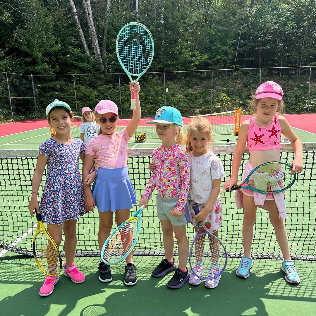 Six young girls standing on a tennis court behind the net, holding tennis rackets, with a wooded background. One girl is raising a teal tennis racket with a dolphin design.