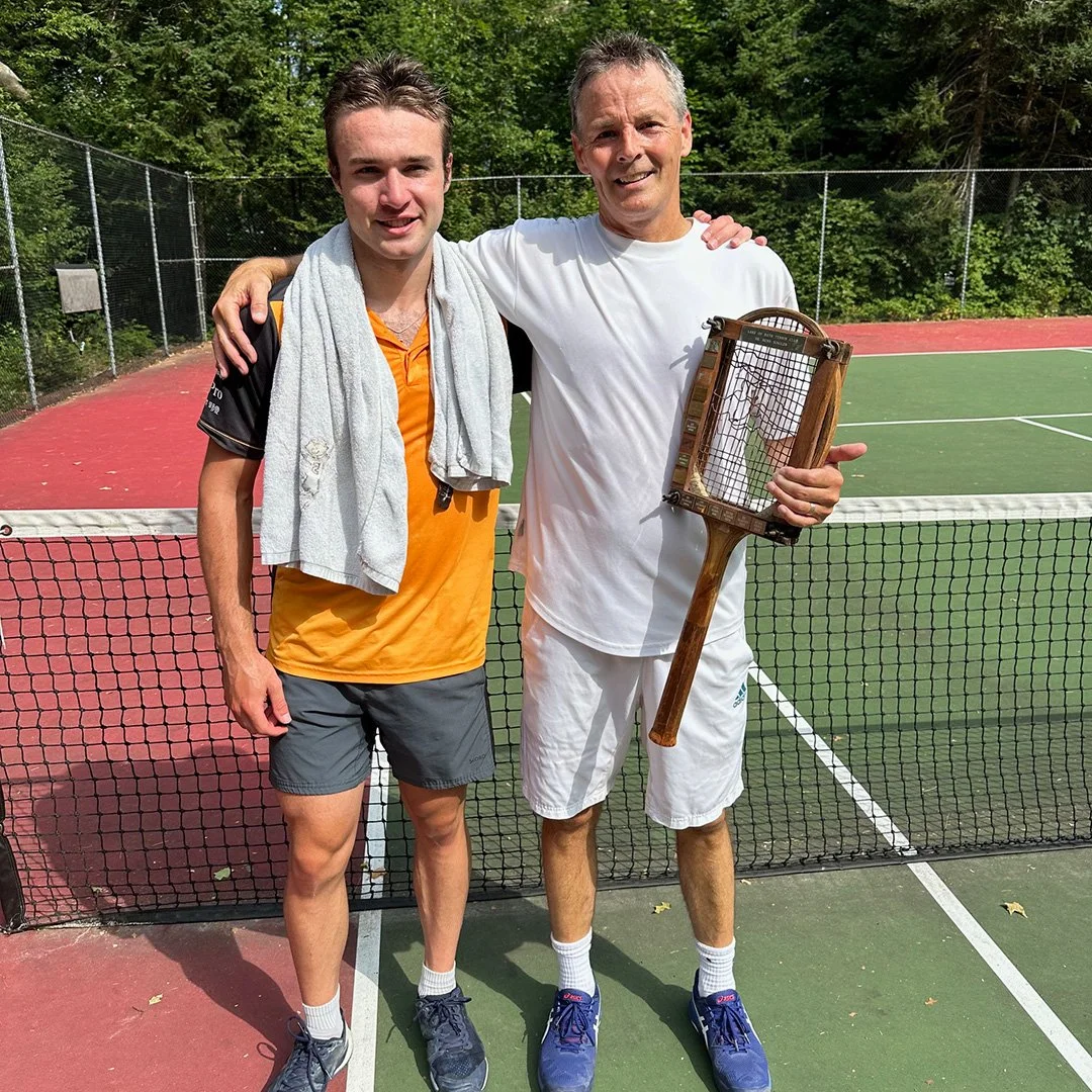 Two men standing on a tennis court, smiling, with one holding a tennis racket. They are dressed in athletic clothing, and the court is surrounded by a fence with trees in the background.