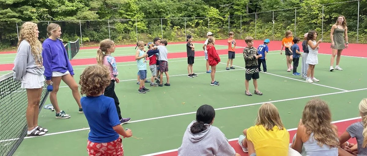Children and adults on an outdoor tennis court. Some children are standing in a line, while others are sitting or standing on the sidelines, with green trees in the background.