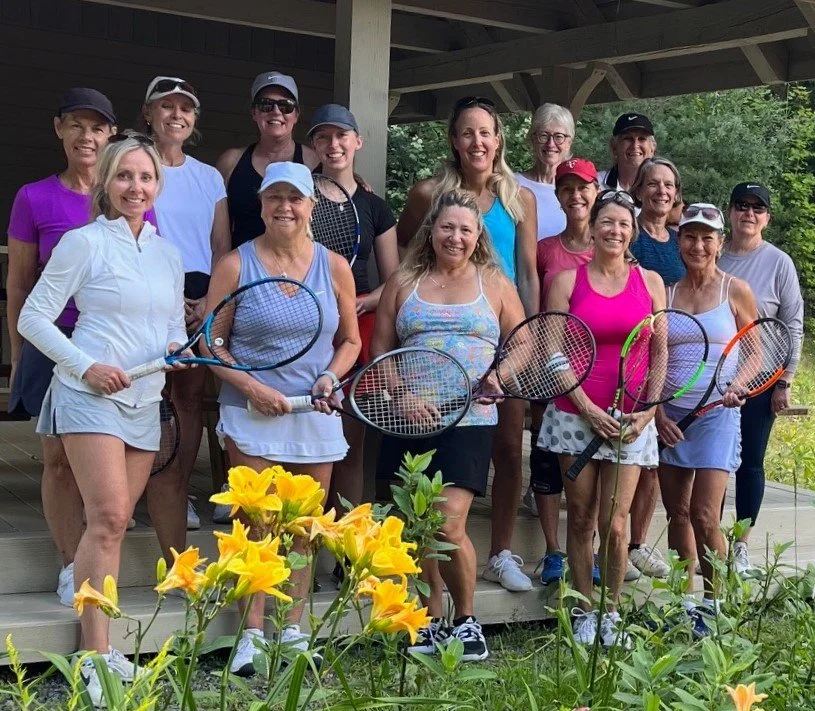 Group of women holding tennis rackets, standing outdoors on a wooden porch, surrounded by yellow flowers and greenery, dressed in sportswear, smiling.