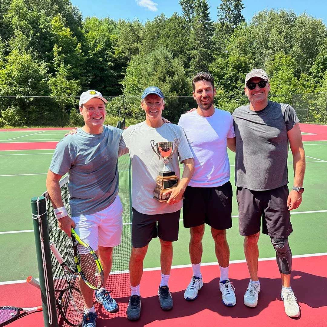 Four men standing on a tennis court, celebrating after a game, with one holding a trophy. They are smiling, dressed in athletic attire, and the background features green trees and a partly cloudy sky.