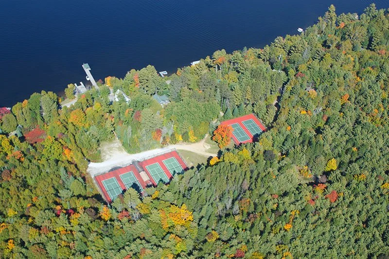 Aerial view of a wooded area with six tennis courts near a lake.