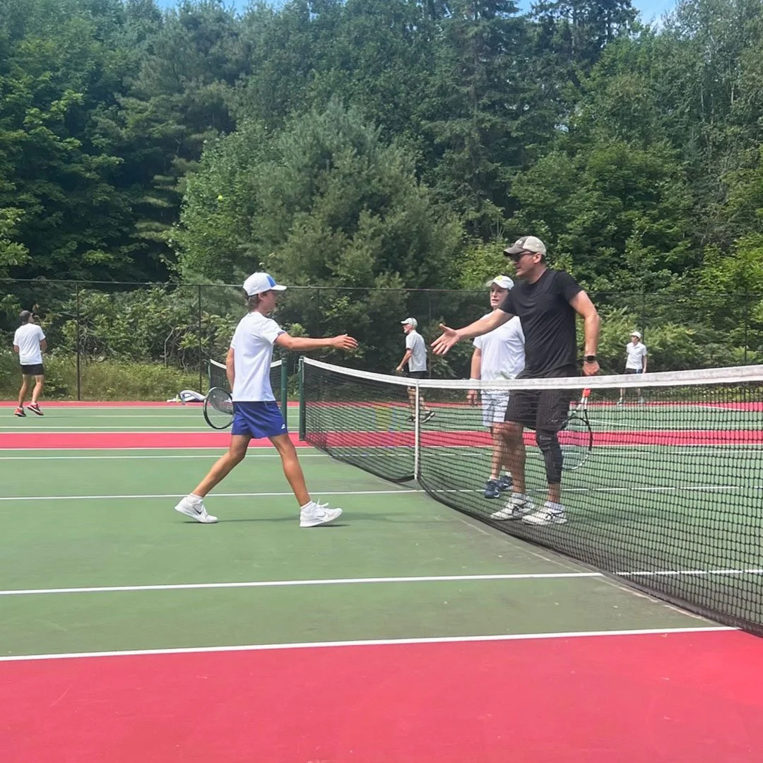 A young tennis player on a green and red outdoor tennis court exchanging a handshake with an adult male after a game, with other players and trees in the background.