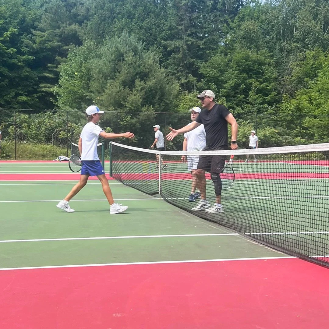 A young boy and a man shake hands on a tennis court after a game, with other players in the background surrounded by trees.