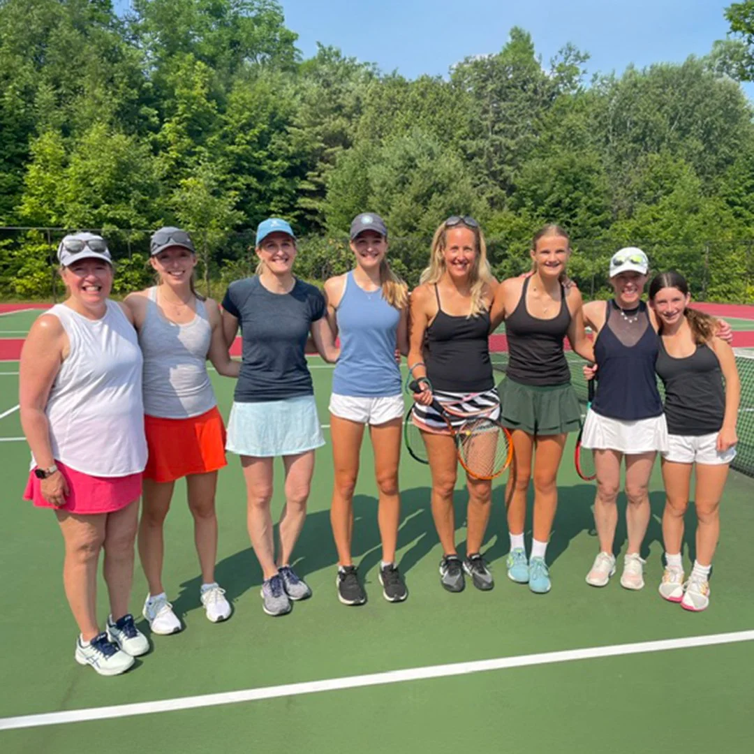 Group of nine women standing arm-in-arm on a tennis court, holding tennis rackets, dressed in athletic attire, with a backdrop of lush green trees under a clear blue sky.