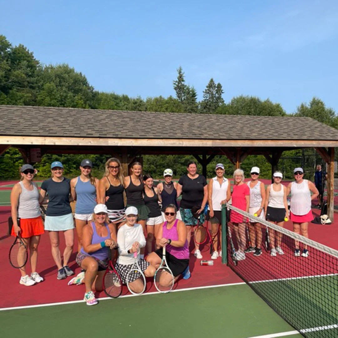 Group of women on a tennis court, holding tennis rackets, posing for a photo, with a pavilion and trees in the background.