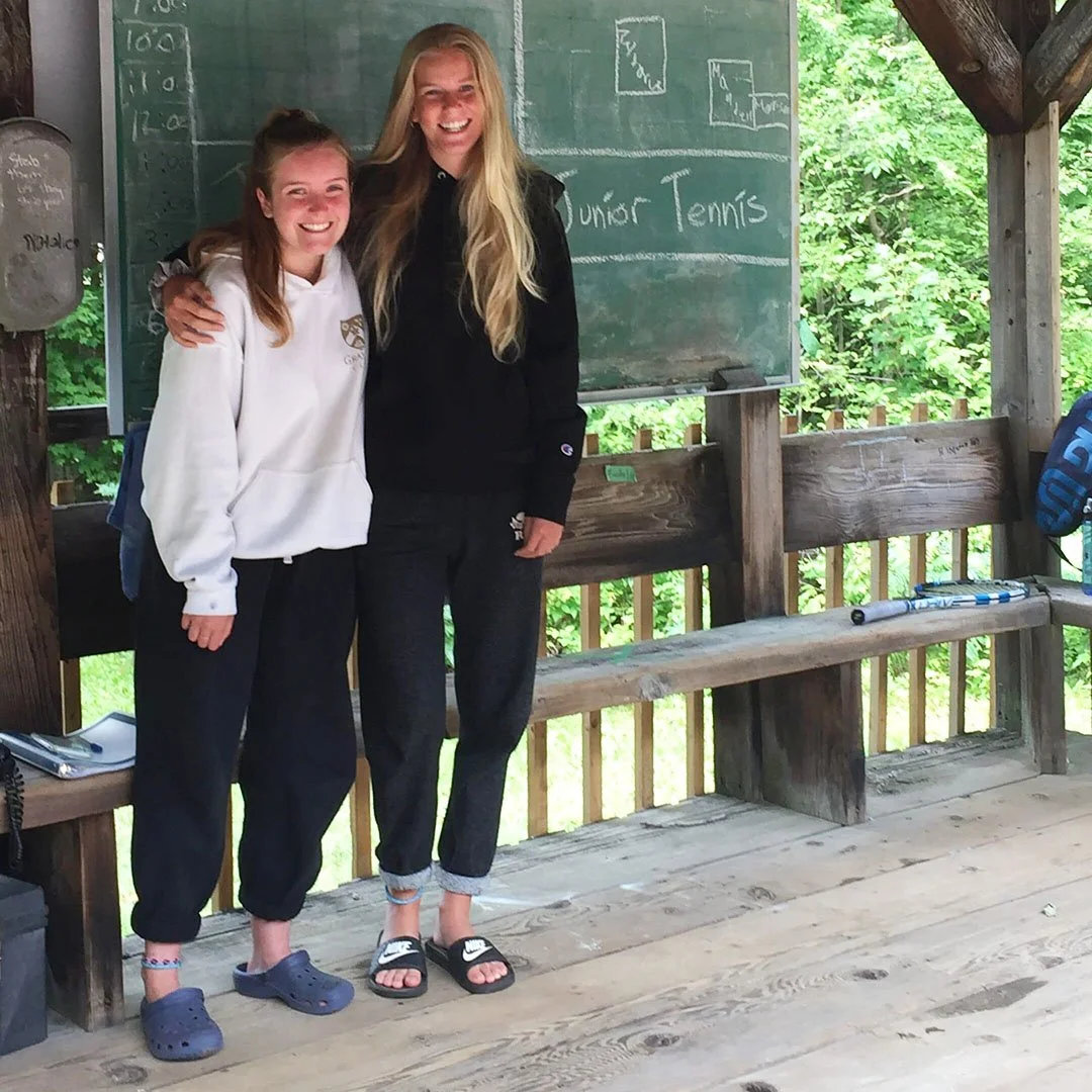 Two teenage girls smiling and posing together on a wooden deck, with a chalkboard behind them that reads "Junior Tennis" and some written schedules, surrounded by trees.