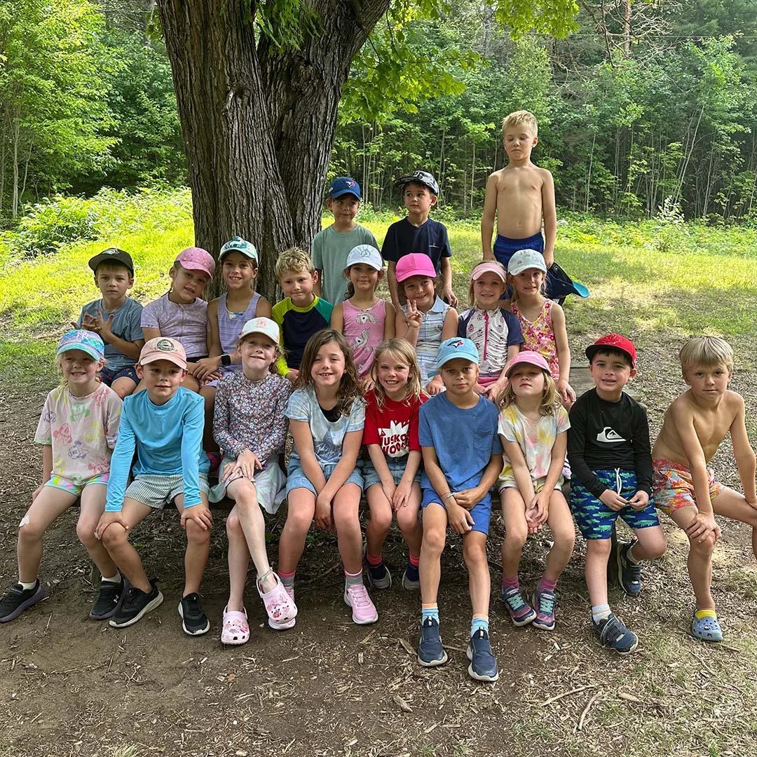 Group of young children sitting and standing outdoors in front of a large tree, some wearing hats, on a sunny day in a lush green forest.