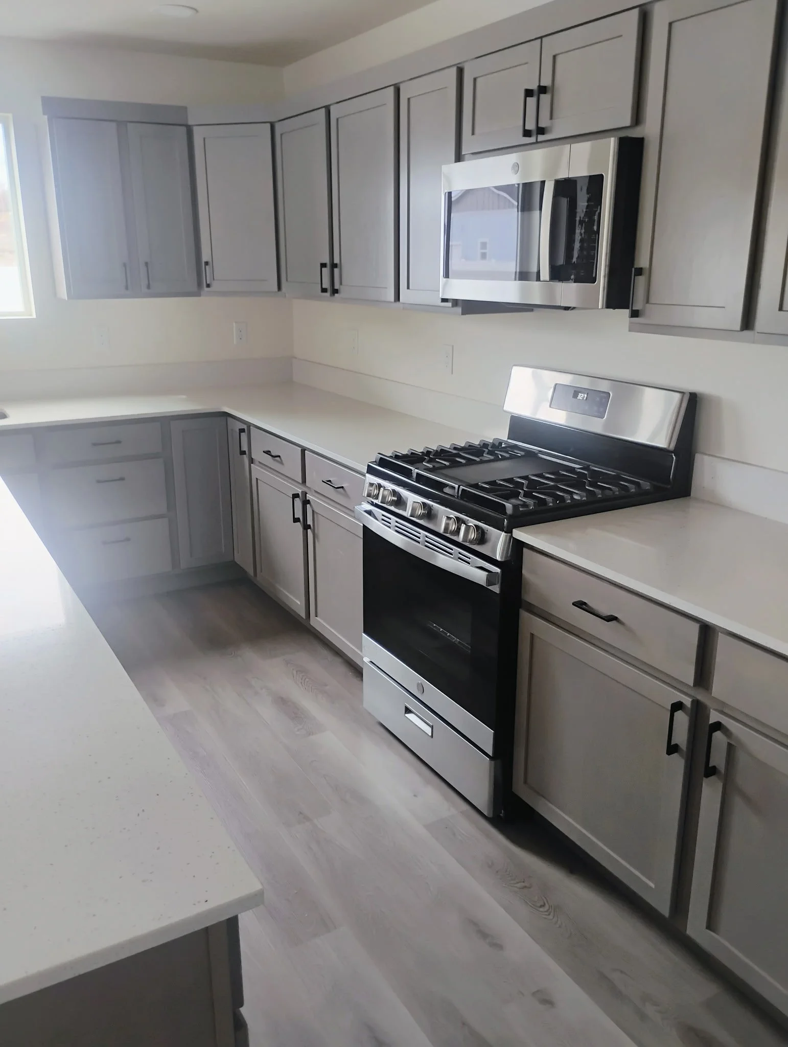 A modern kitchen with gray cabinets, a black and stainless steel stove, and a stainless steel microwave above the stove. The kitchen has light-colored wood flooring and white countertops. There is a window on the left side letting in natural light.