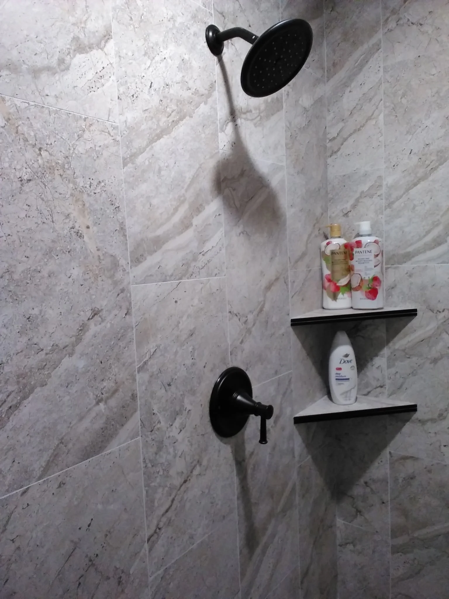 A shower area with marble tiles and a black shower head. There are two bottles of soap or shampoo on a black shelf, and a bottle of Dove body wash on the lower shelf.