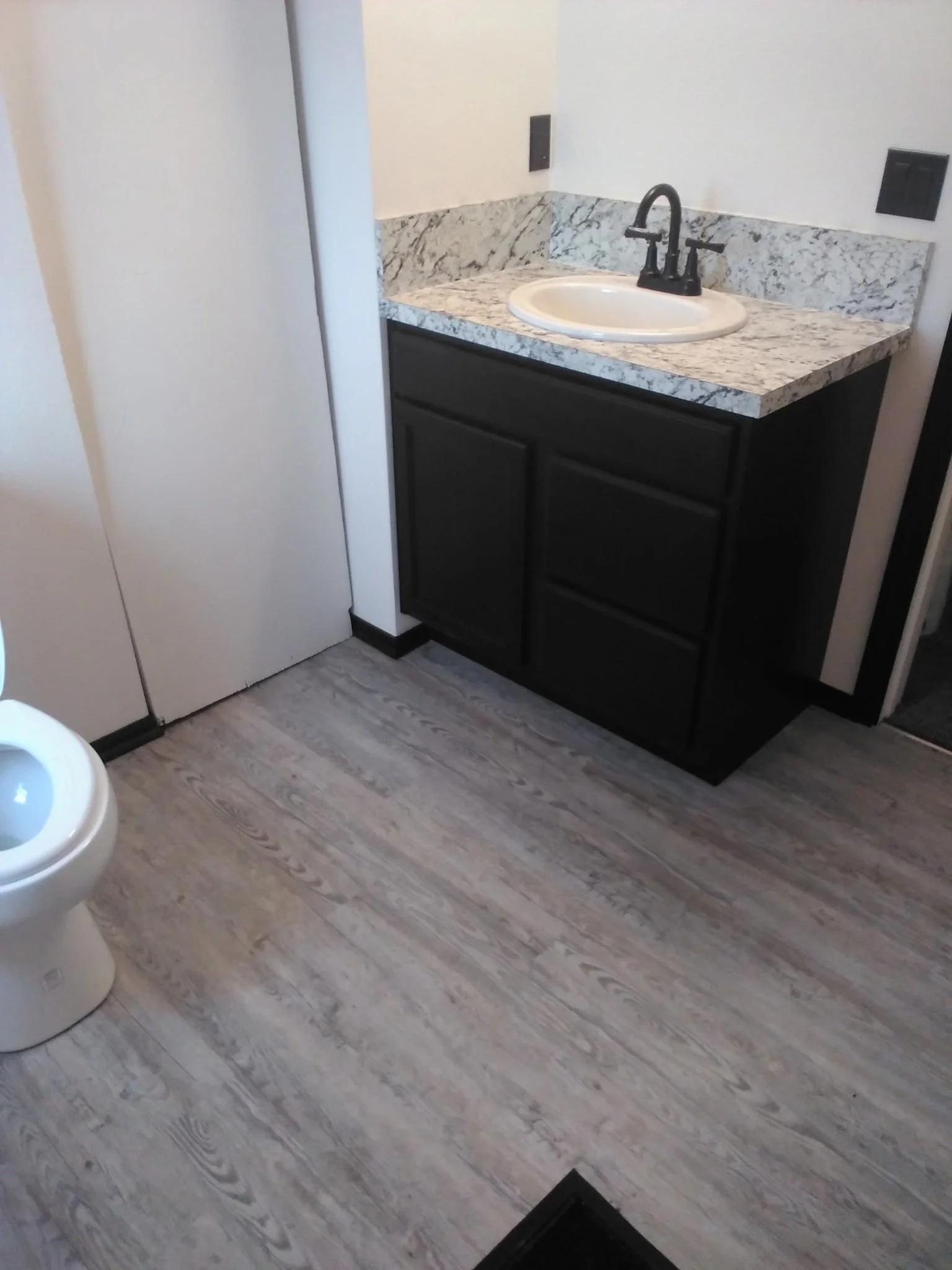 A bathroom with a toilet and a sink with a marble countertop and black cabinetry. The room has wood-look flooring and white walls with a black electrical outlet visible.