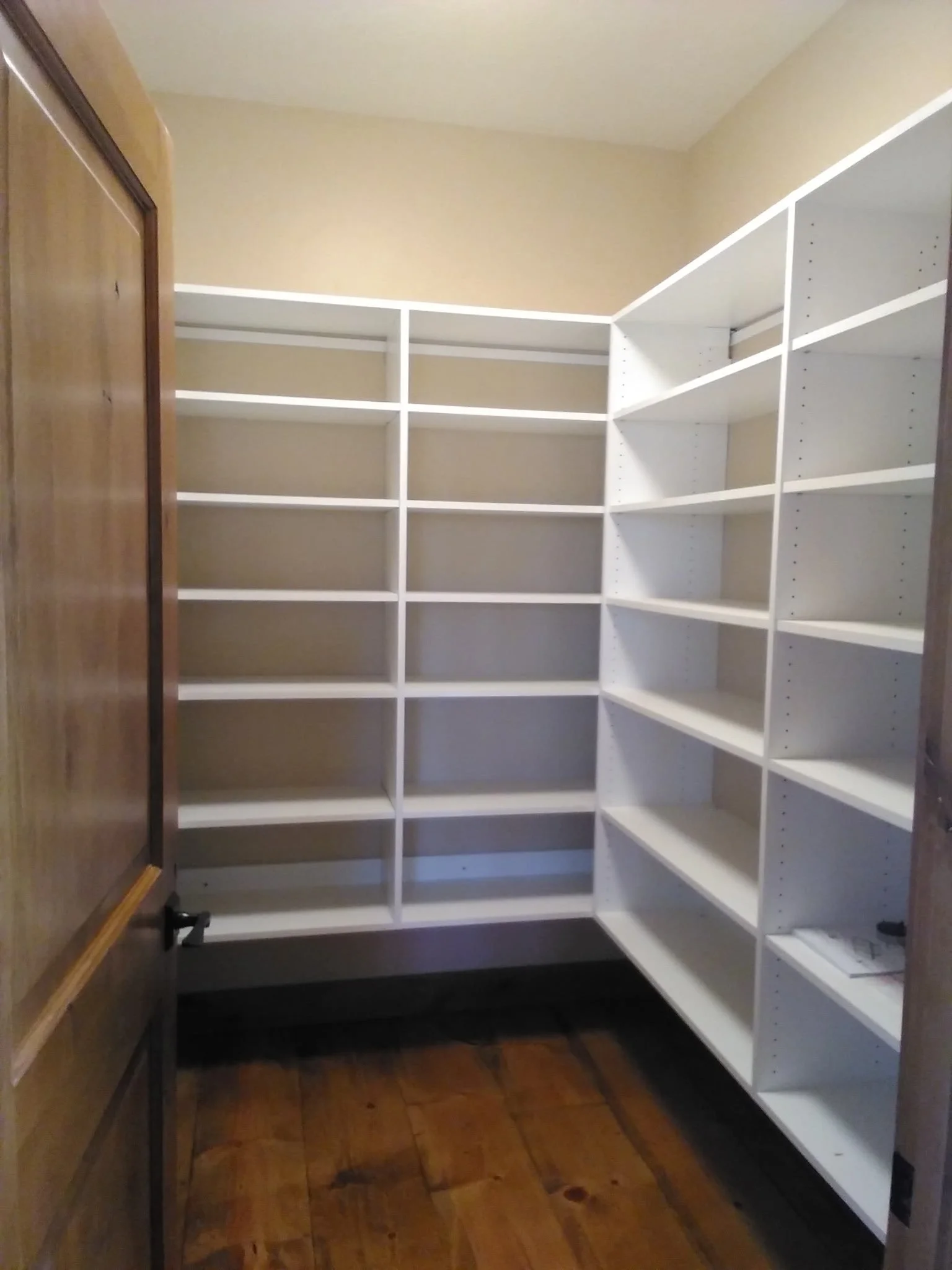 Empty white built-in corner shelves in a small closet with beige walls and hardwood floors.