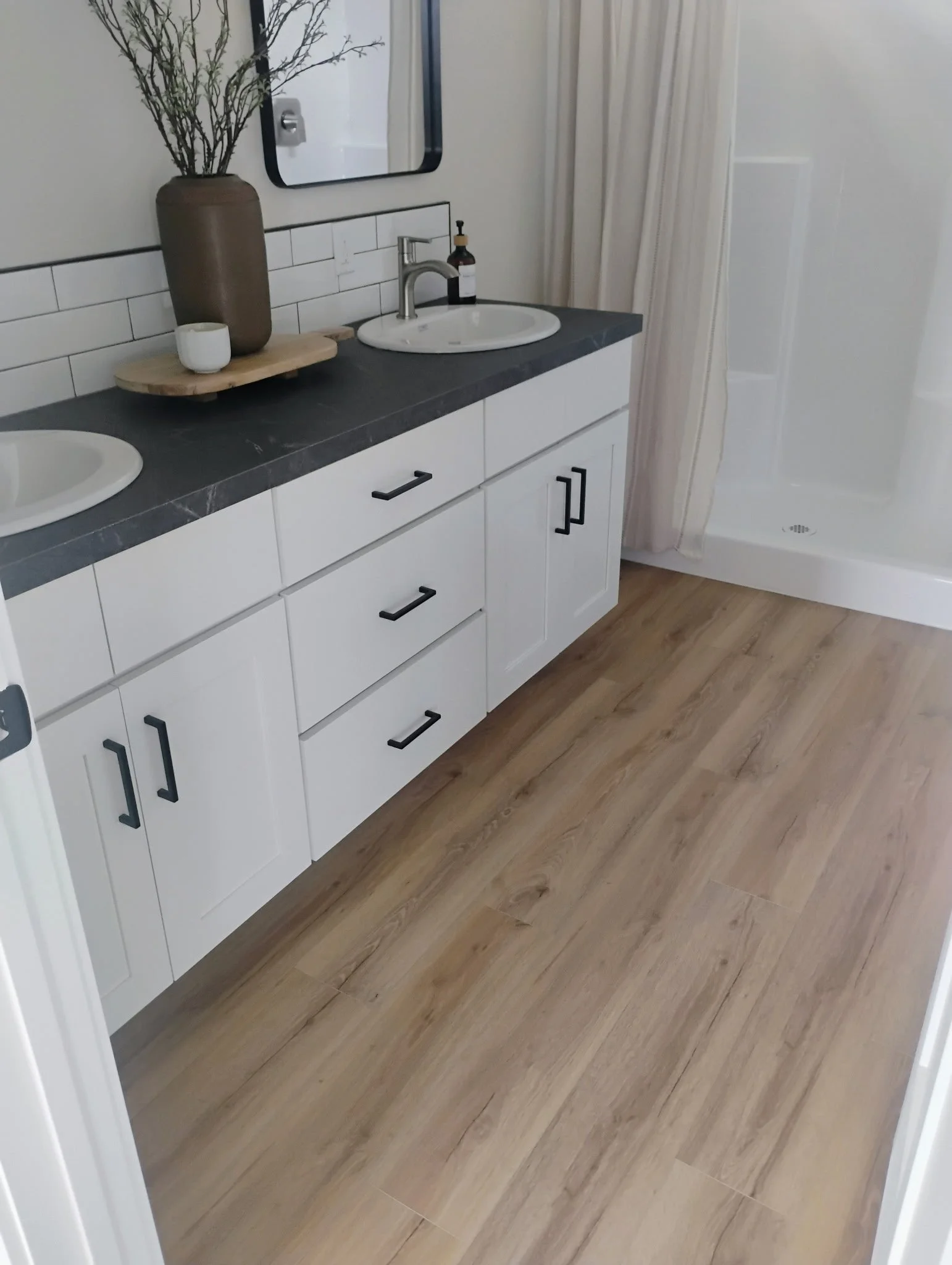 A bathroom vanity with a dark countertop, white cabinets with black handles, and a sink. Decor includes a tall brown vase with decorative branches, a small white bowl, and a brown soap dispenser. A mirror hangs above the vanity, and a shower with a c