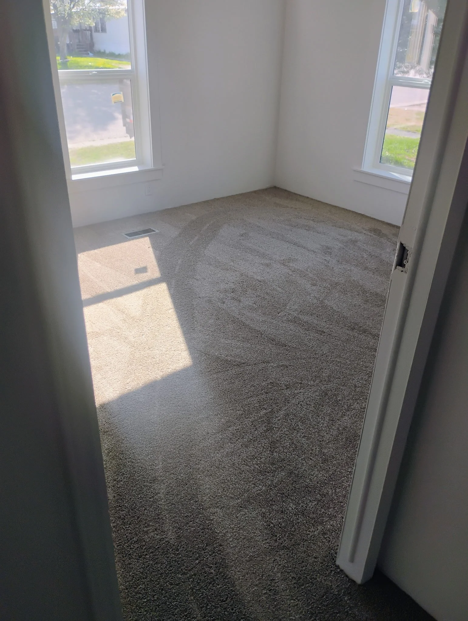 Empty room with beige carpet, two large windows allowing sunlight, white walls, and a partially open door.