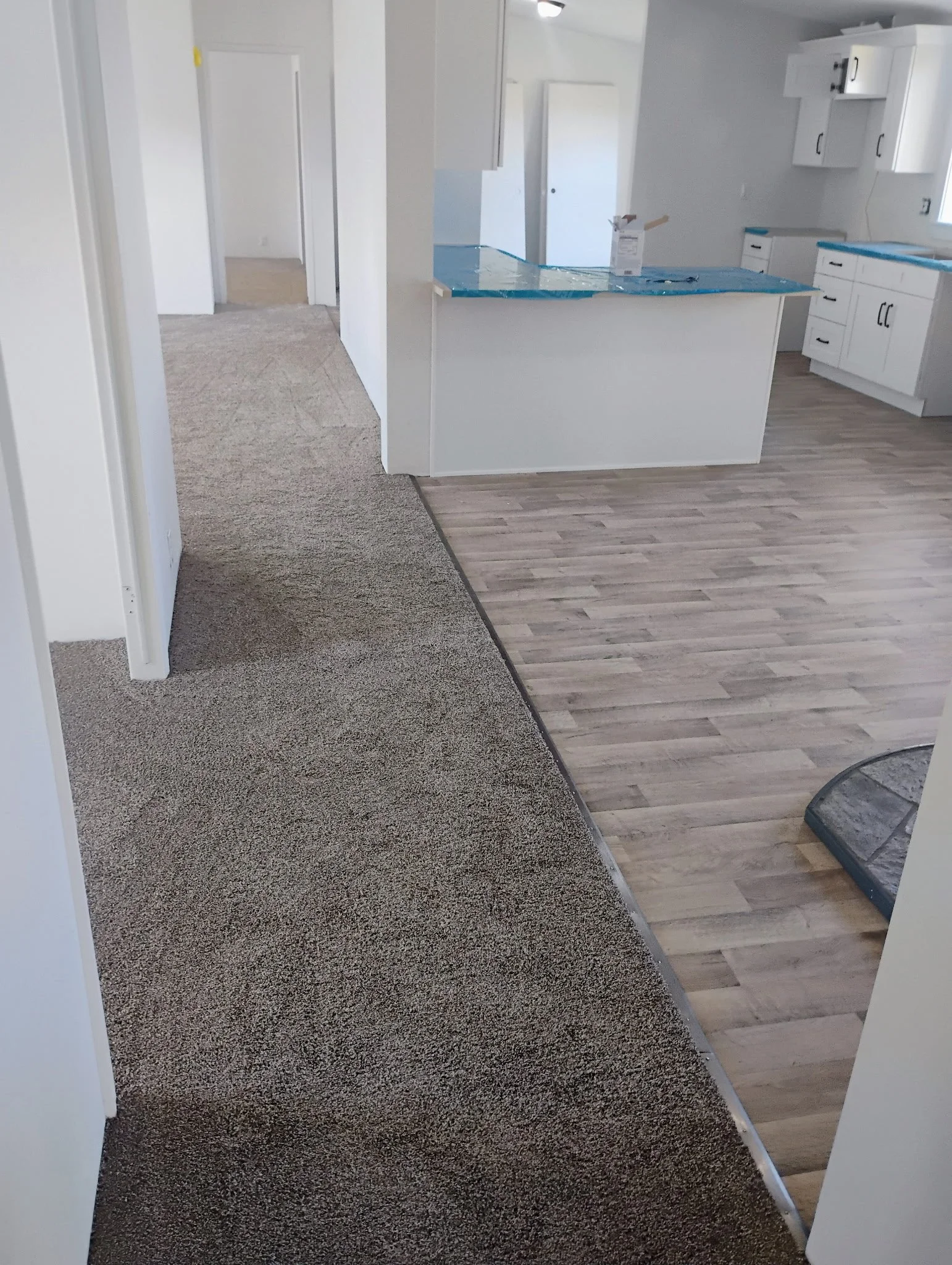A room under renovation with the kitchen area on the right and a carpeted area on the left. The kitchen has white cabinets and a partially installed countertop covered with blue protective plastic. The flooring alternates between carpet and wood lami