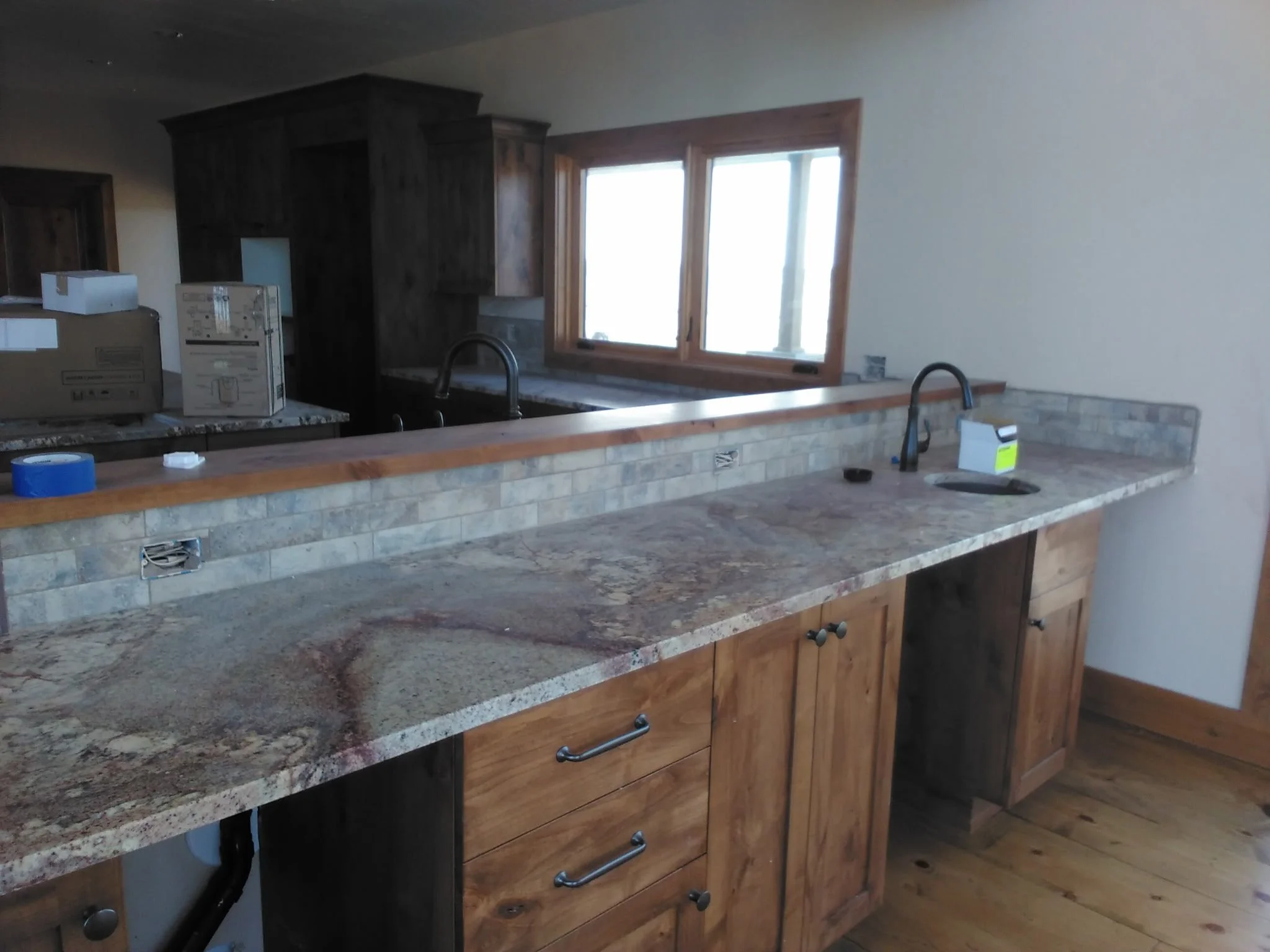 A kitchen with a granite countertop, wooden cabinets, and a window above the sink. Various boxes and packing materials are on the counter, indicating ongoing moving or remodeling.