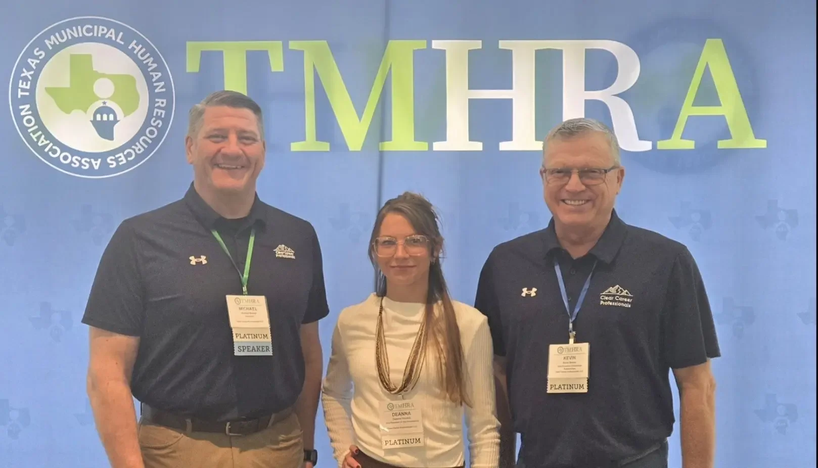 Three people standing in front of a blue background with the logo of the Texas Municipal Human Resources Association (TMHRA). The two men are wearing black polo shirts with TMHRA logos and name tags that identify them as platinum speakers, while the woman in the middle is wearing glasses, a cream-colored top, and a statement necklace, also with a platinum badge.