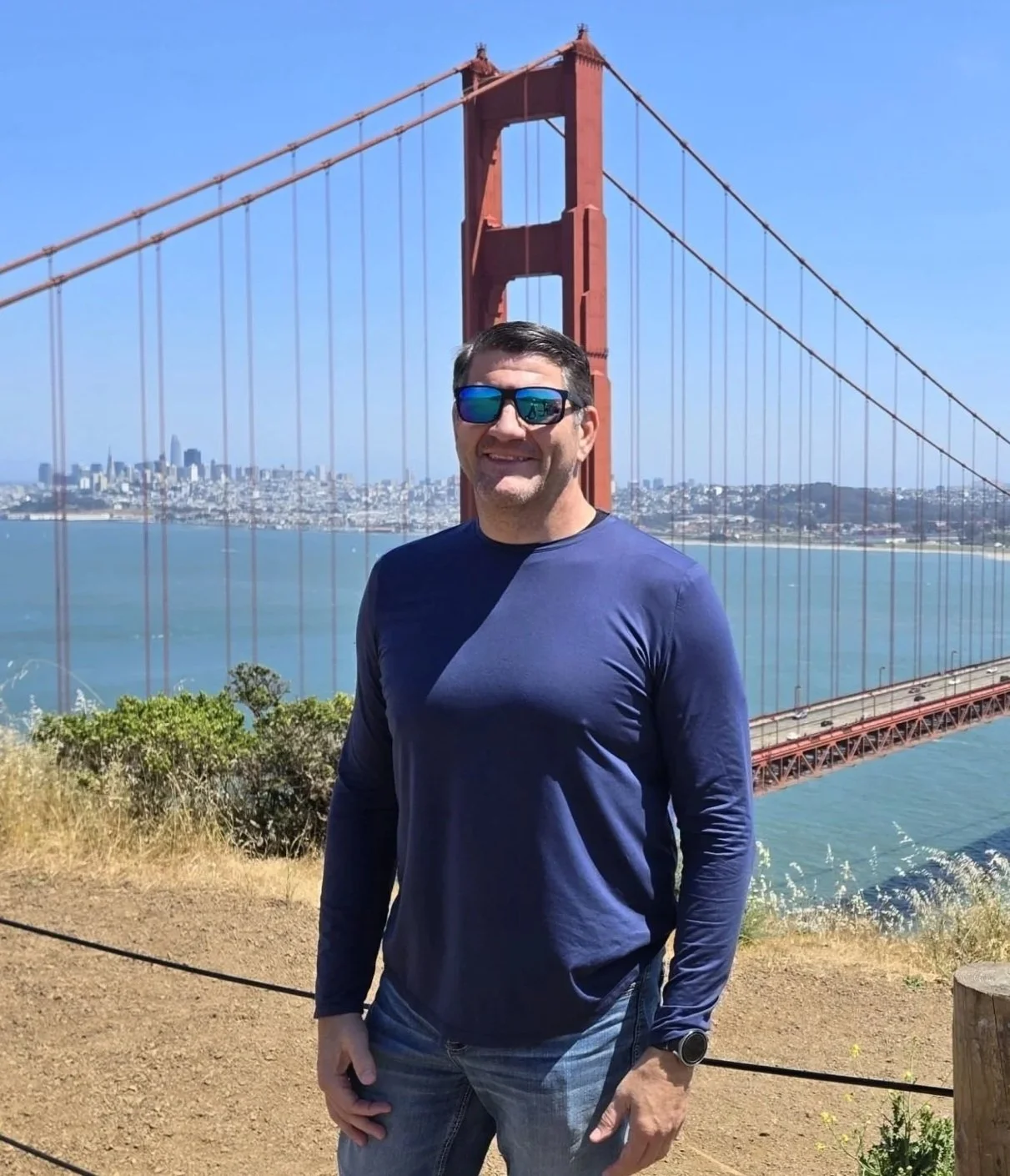 A man wearing sunglasses and a blue long-sleeve shirt standing outdoors with the Golden Gate Bridge in the background on a clear day.