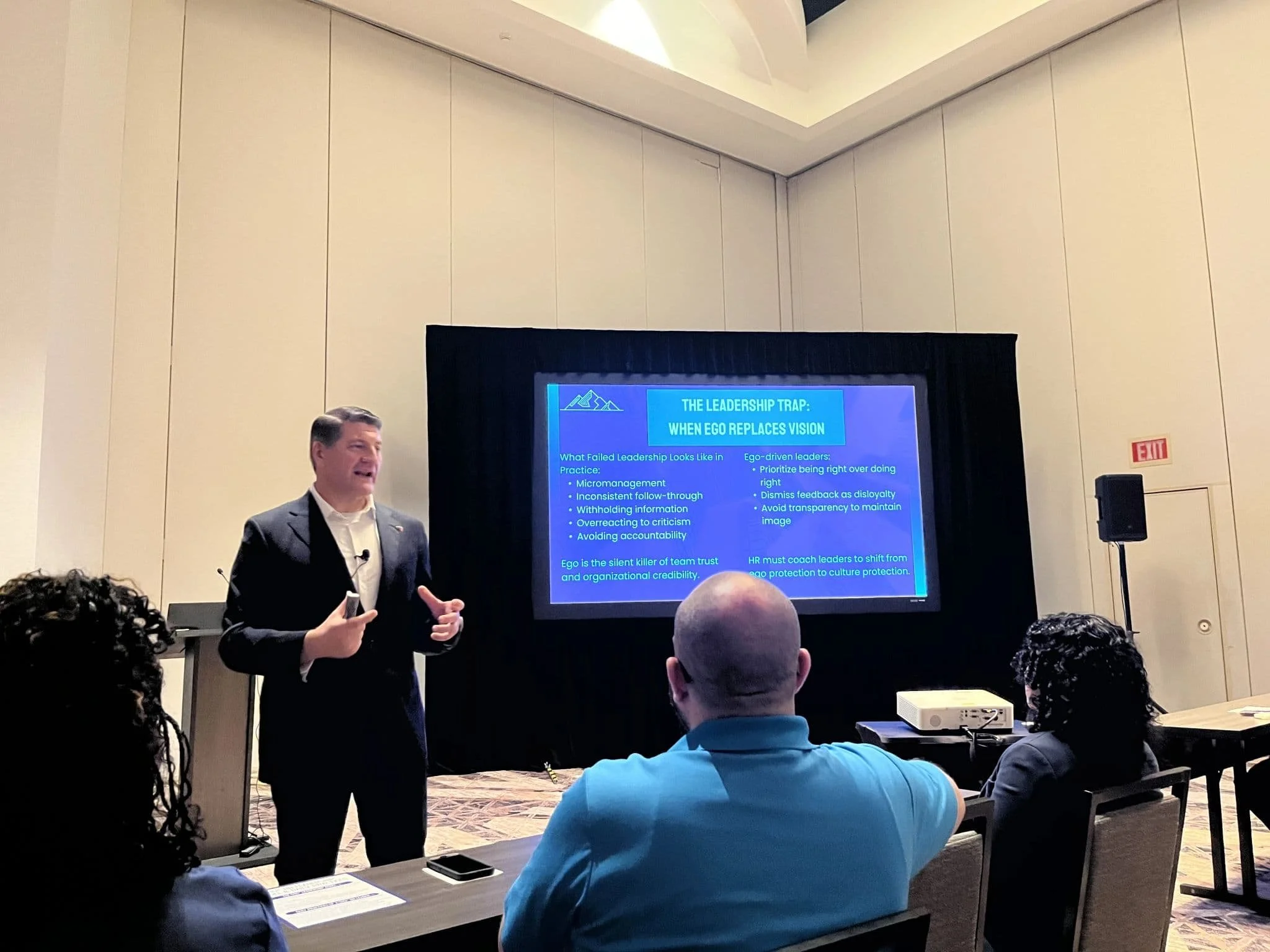 Michael Boese in a suit giving a presentation in a conference room with a large screen displaying a slide titled 'The Leadership Trap: When Ego Replaces Vision.' There are audience members seated at tables, some taking notes.