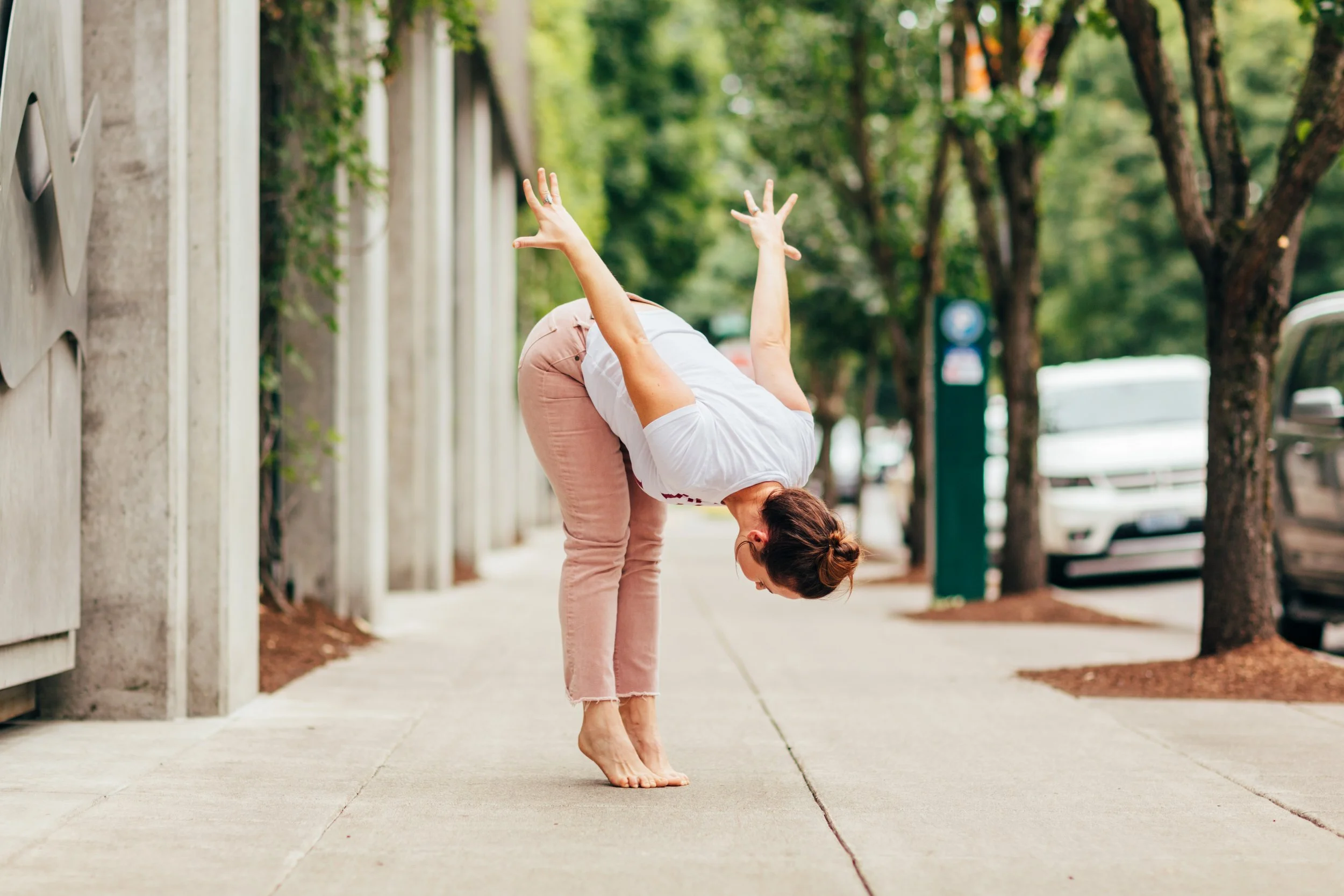 Woman in white T-shirt and pink jeans practicing yoga on sidewalk in a tree-lined urban area.