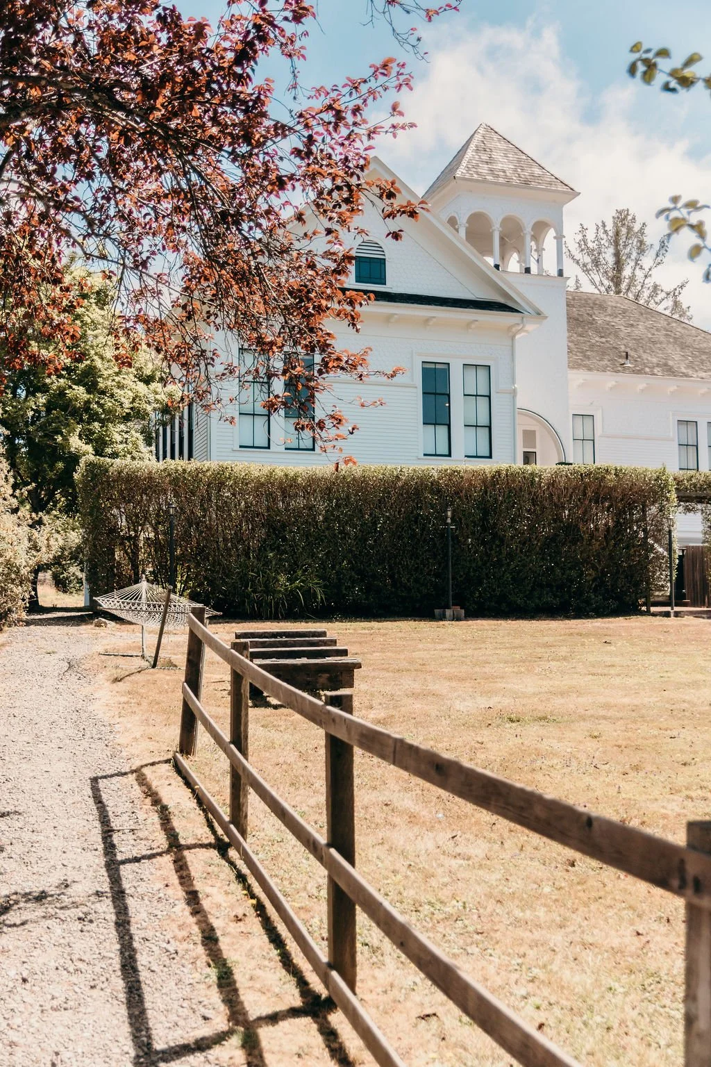 A white historic schoolhouse with a tower and arched windows, a brown shingled roof, surrounded by a hedge, a gravel walkway with wooden fencing, grass yard, trees with reddish leaves, and a blue sky with clouds.