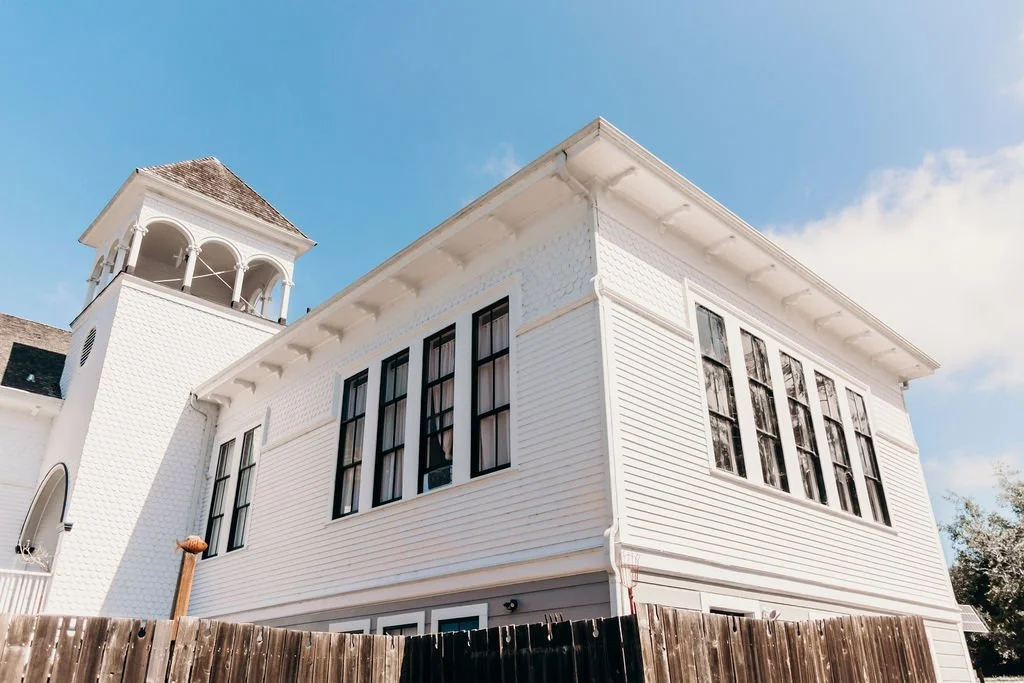 A white multi-story house with black window frames and a tower-like structure with a shingled roof, set against a blue sky with some clouds.