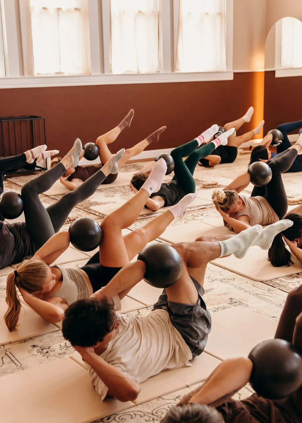 People participating in a fitness class doing core exercises with a small Pilates ball, lying on mats in a studio with natural light.