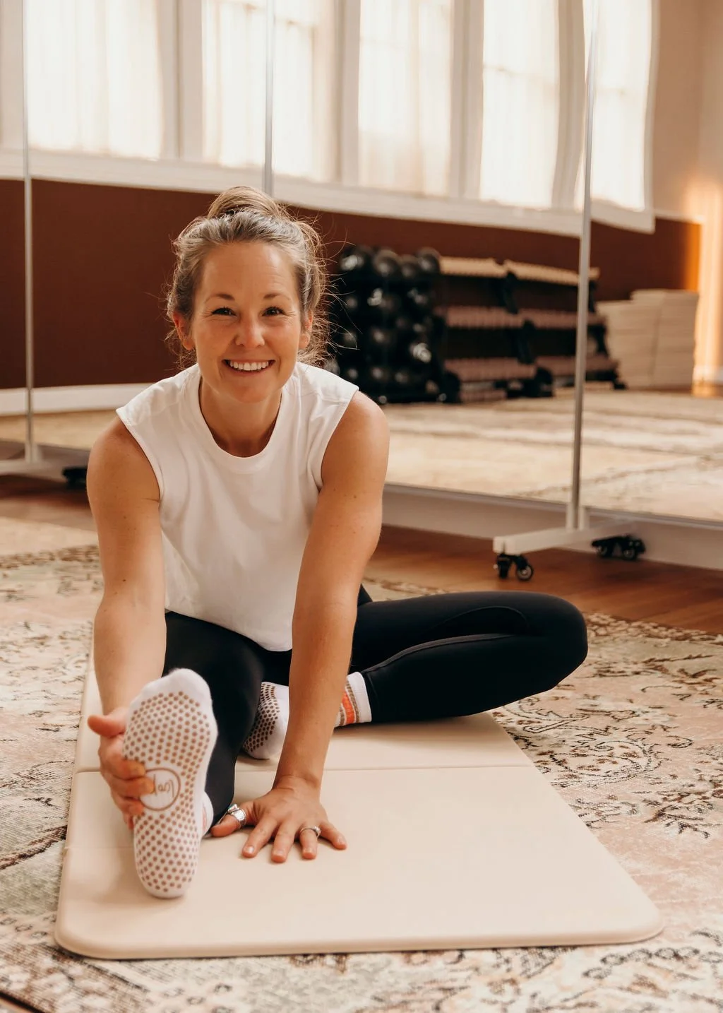 A woman sitting on a Stakt Pilates mat in a fitness room, stretching her leg while smiling at the camera.
