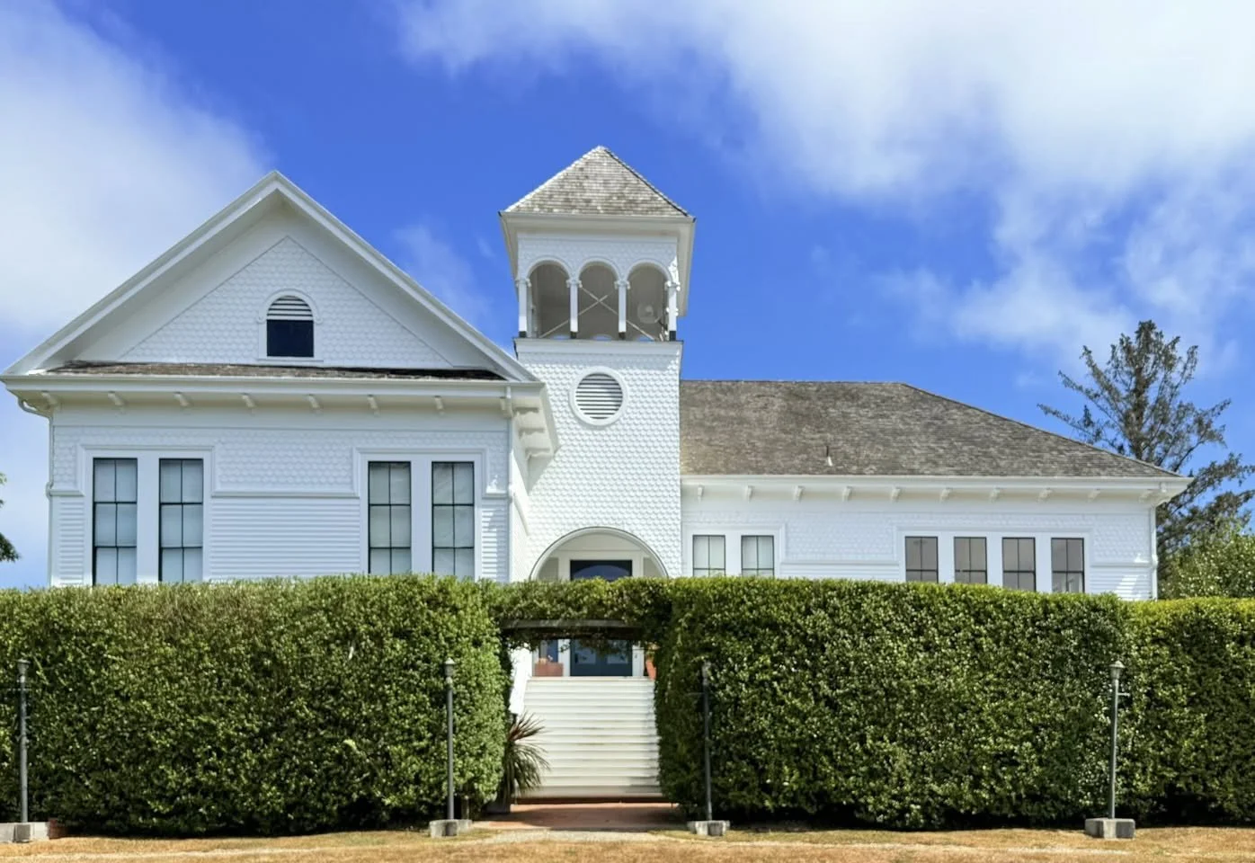 White historic house with a tower, surrounded by a neatly trimmed hedge, set against a blue sky with clouds.