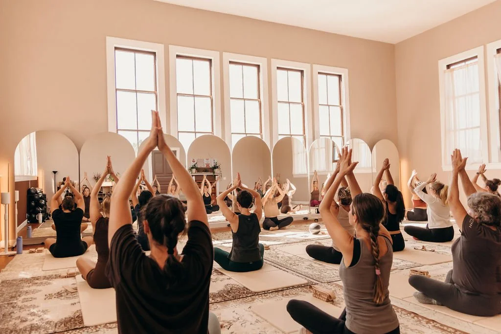 A group of people participating in a barre or Pilates class, sitting on mats with their arms raised and hands pressed together in a prayer position, in a bright studio with large windows.