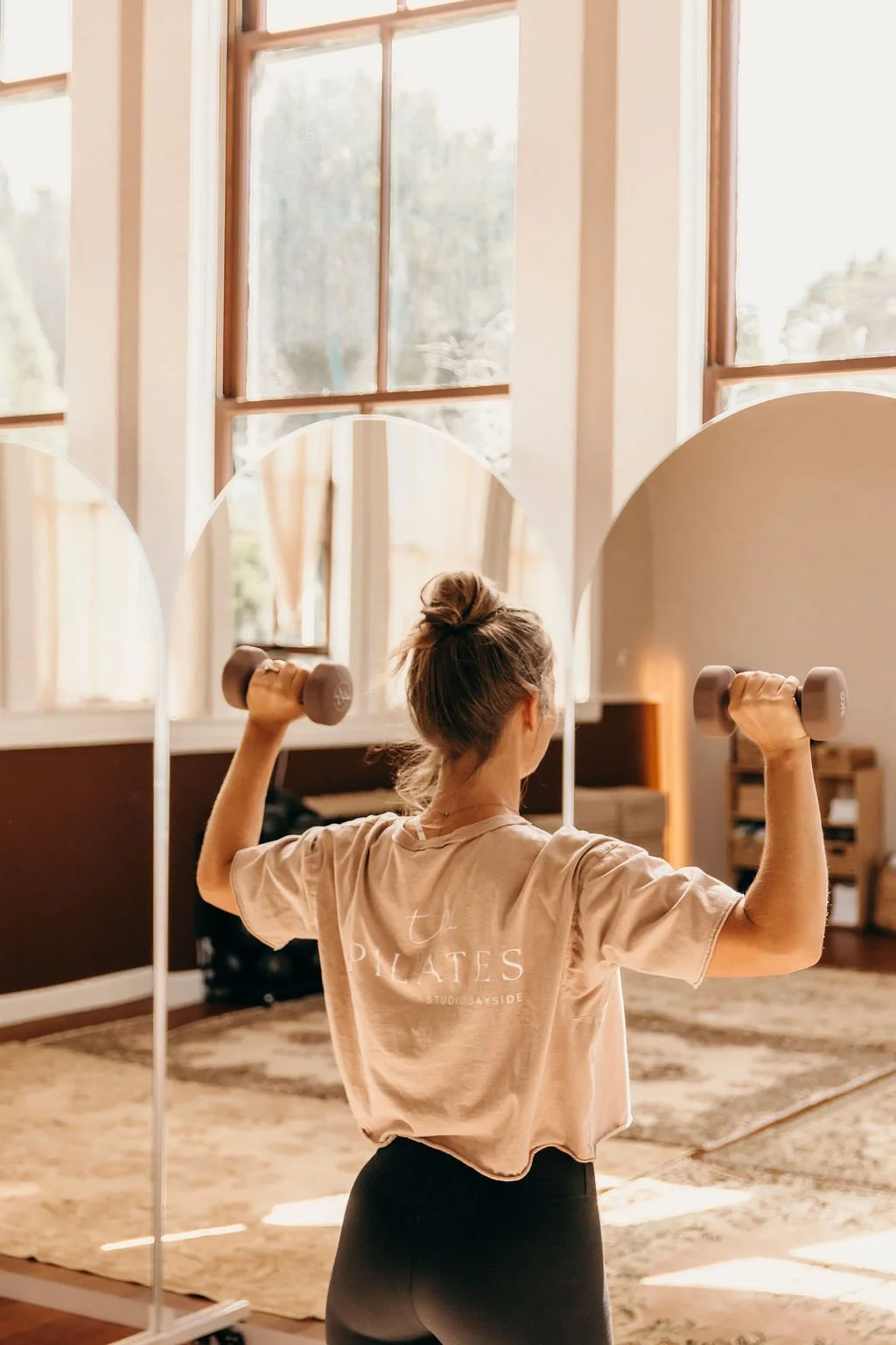 A woman with a bun hairstyle wearing a beige t-shirt with 'Pilates' printed on the back, lifting dumbbells in front of a mirror in a room with large windows, a rug, and wooden furniture.