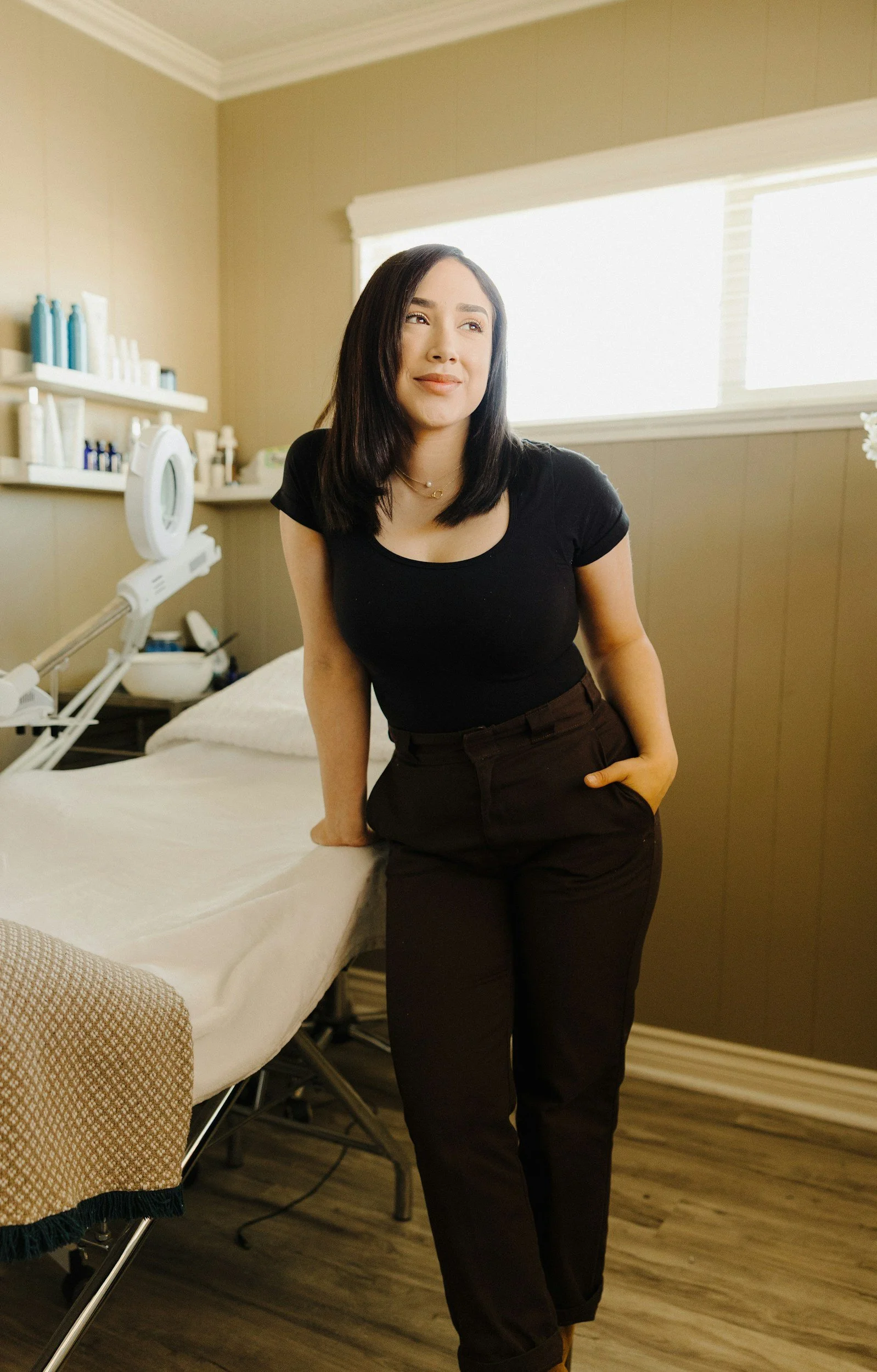 A woman with dark hair wearing a black t-shirt and brown pants leaning on a hospital bed in a room with neutral-colored walls and medical supplies on shelves.