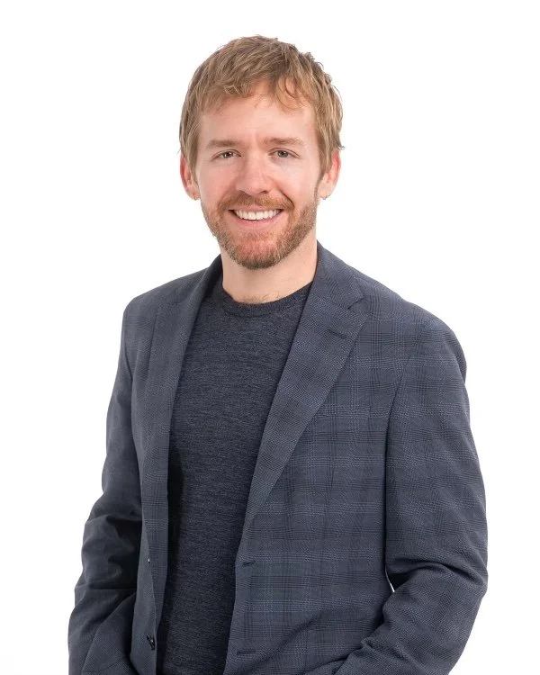 A man with light brown curly hair and a beard, smiling, wearing a dark gray blazer over a crew neck shirt, against a white background.
