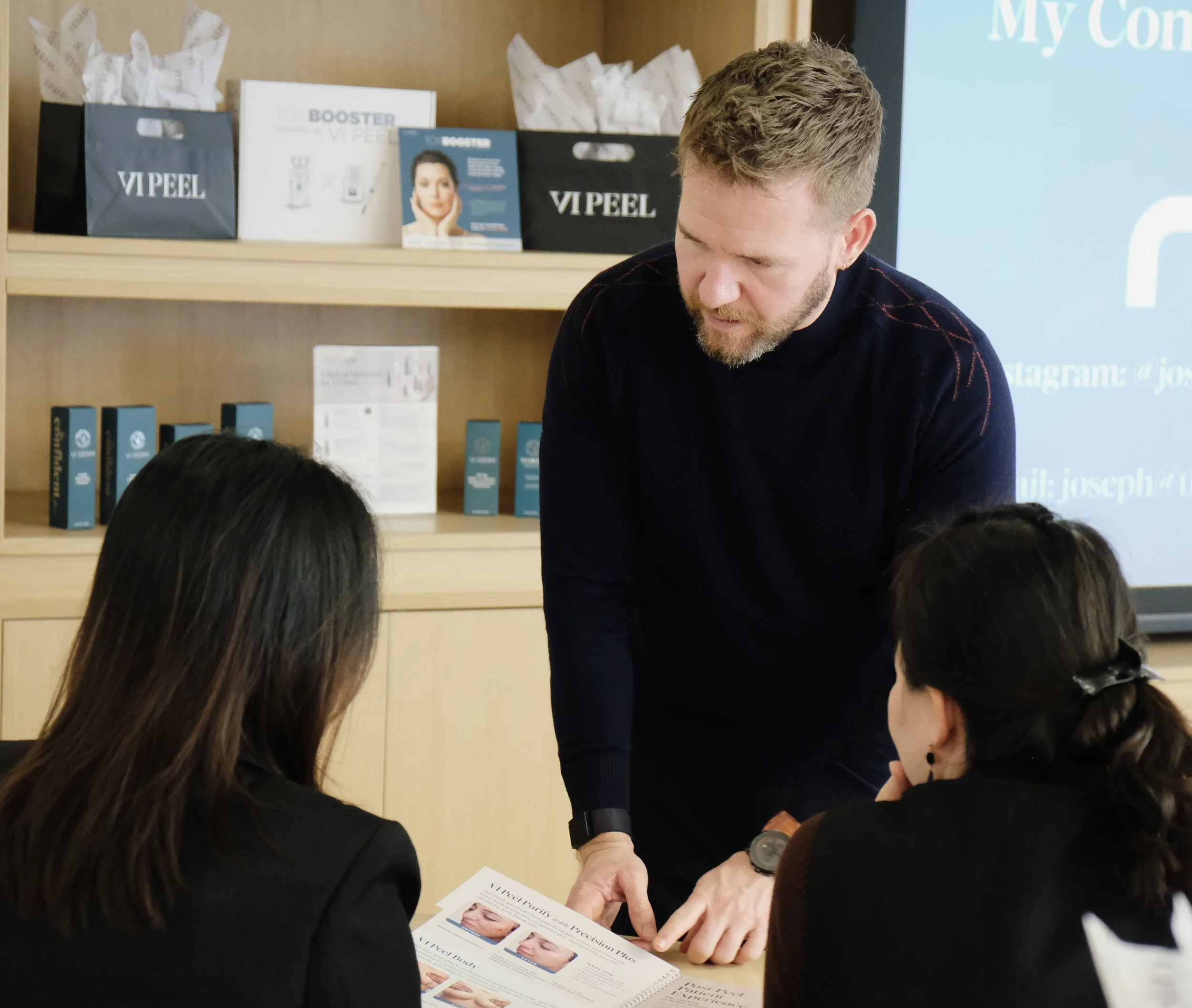 A man and two women in a room with a wooden shelf and promotional materials, engaged in a discussion or presentation.