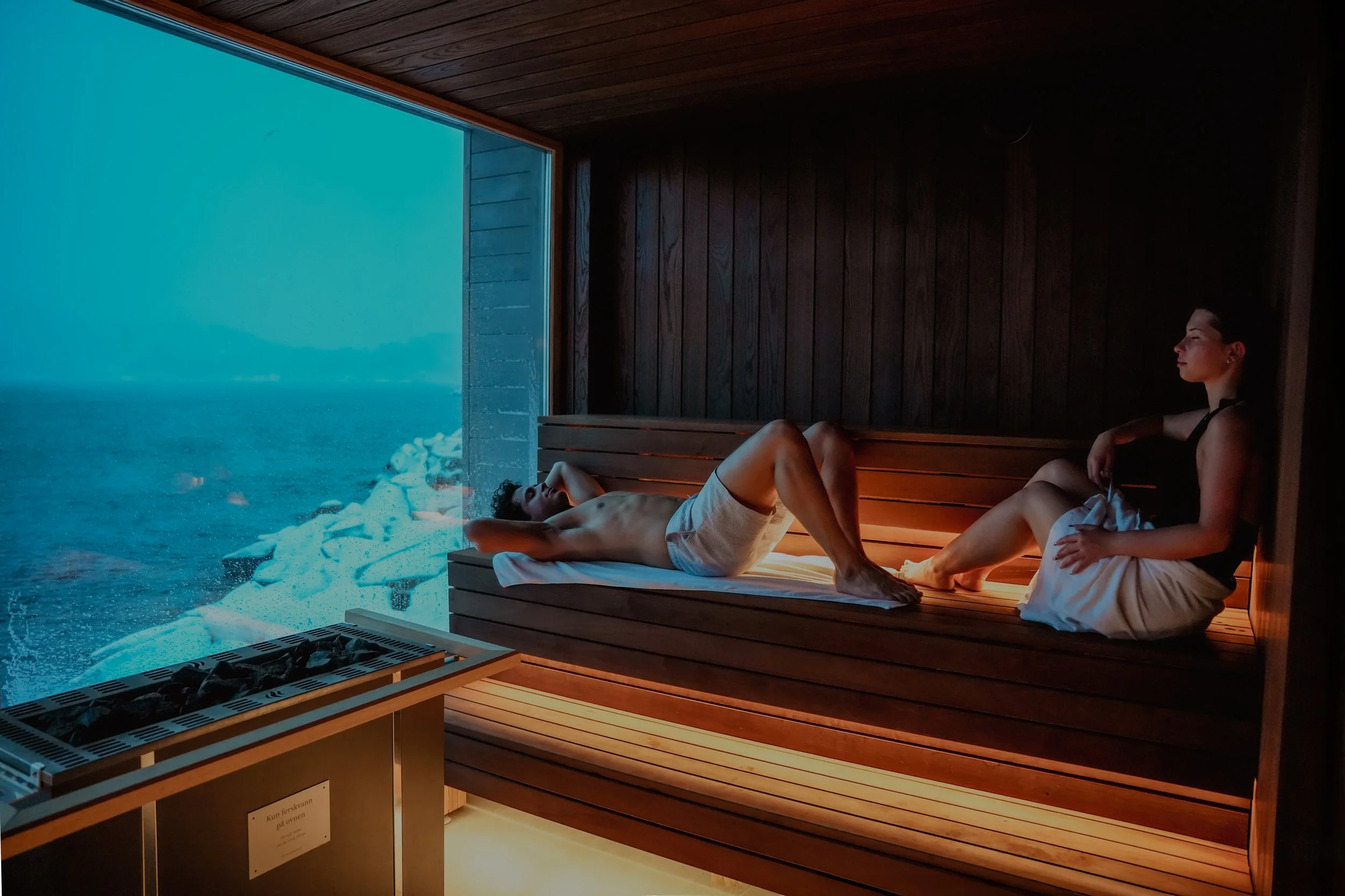 A man and a woman relax in a wood-paneled sauna overlooking the ocean through a large window.