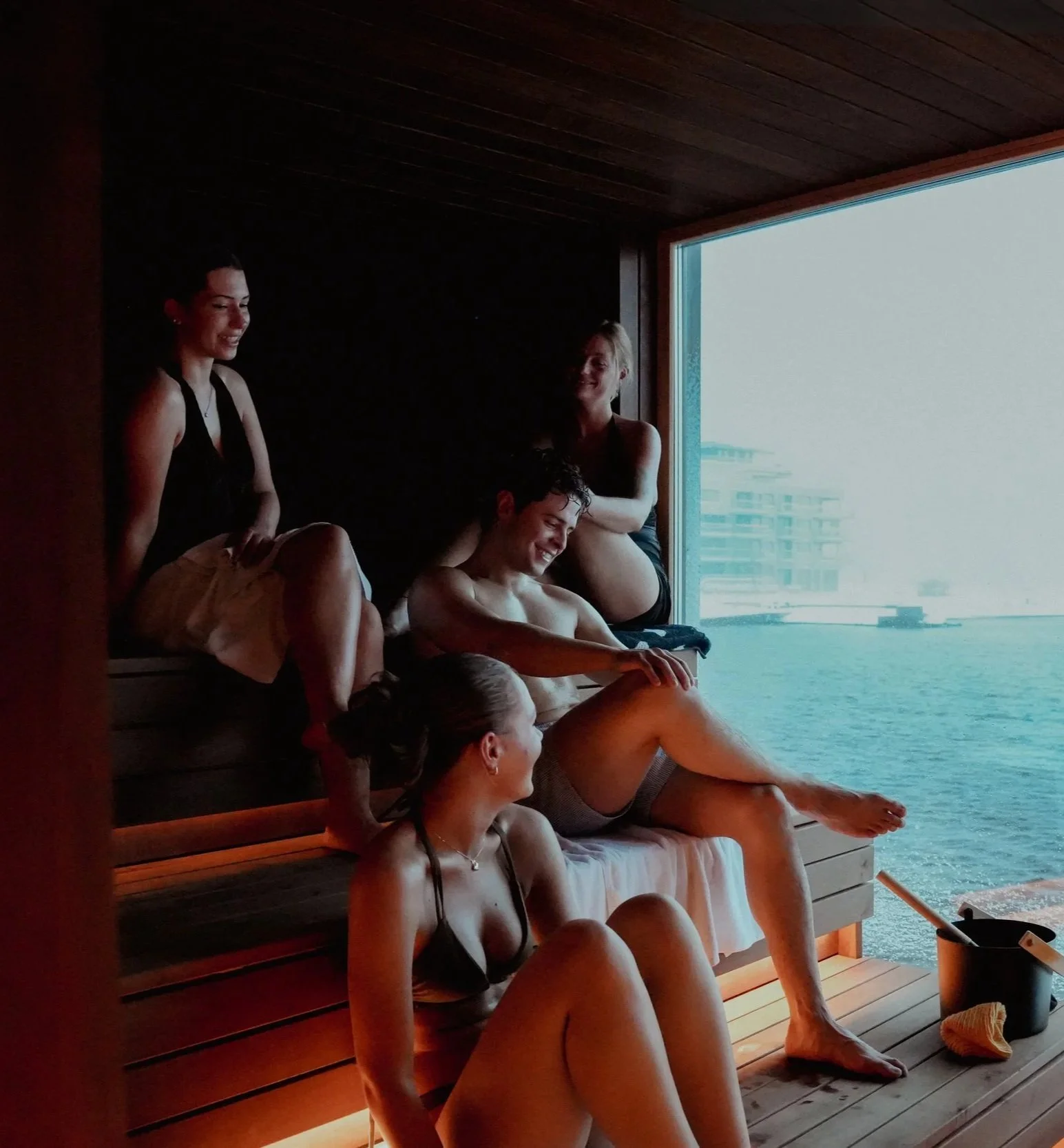 A group of five young adults relaxing inside a wooden sauna with a large window overlooking water and buildings outside.