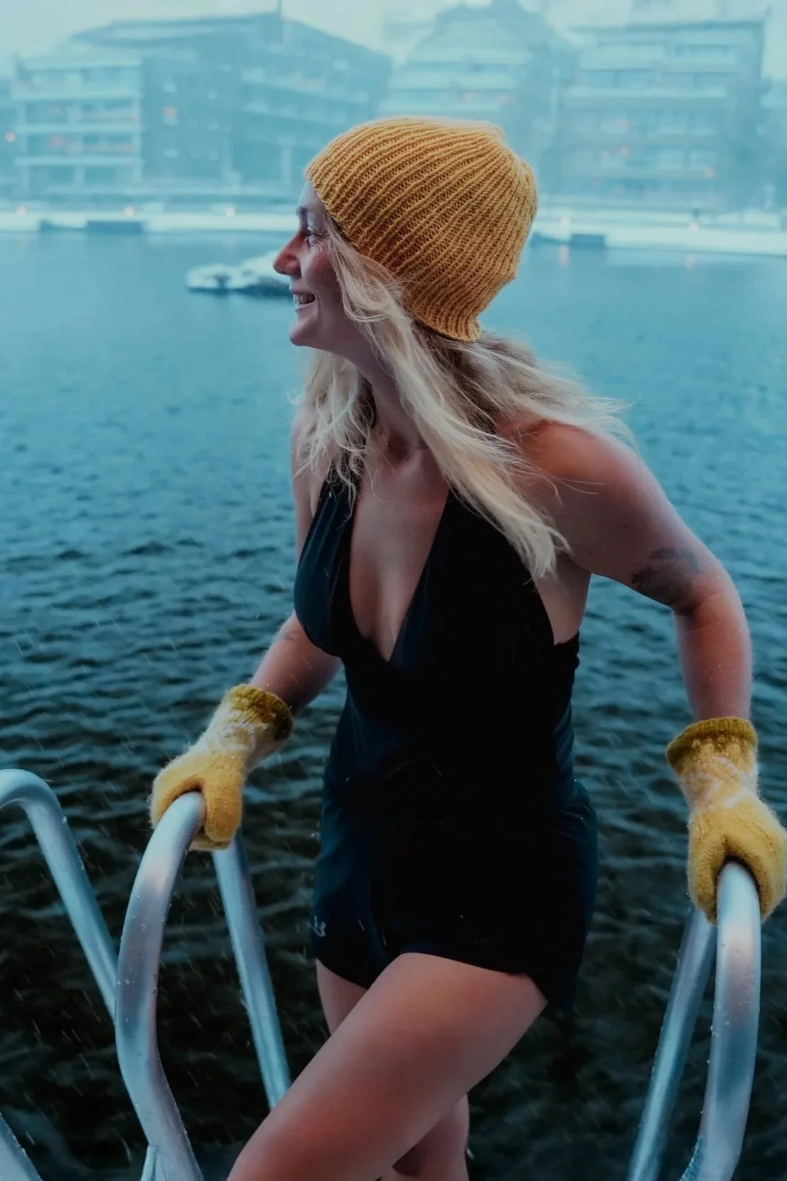 A woman wearing a yellow knit beanie and yellow gloves climbing out of a body of water, with a cityscape with modern buildings in the background.